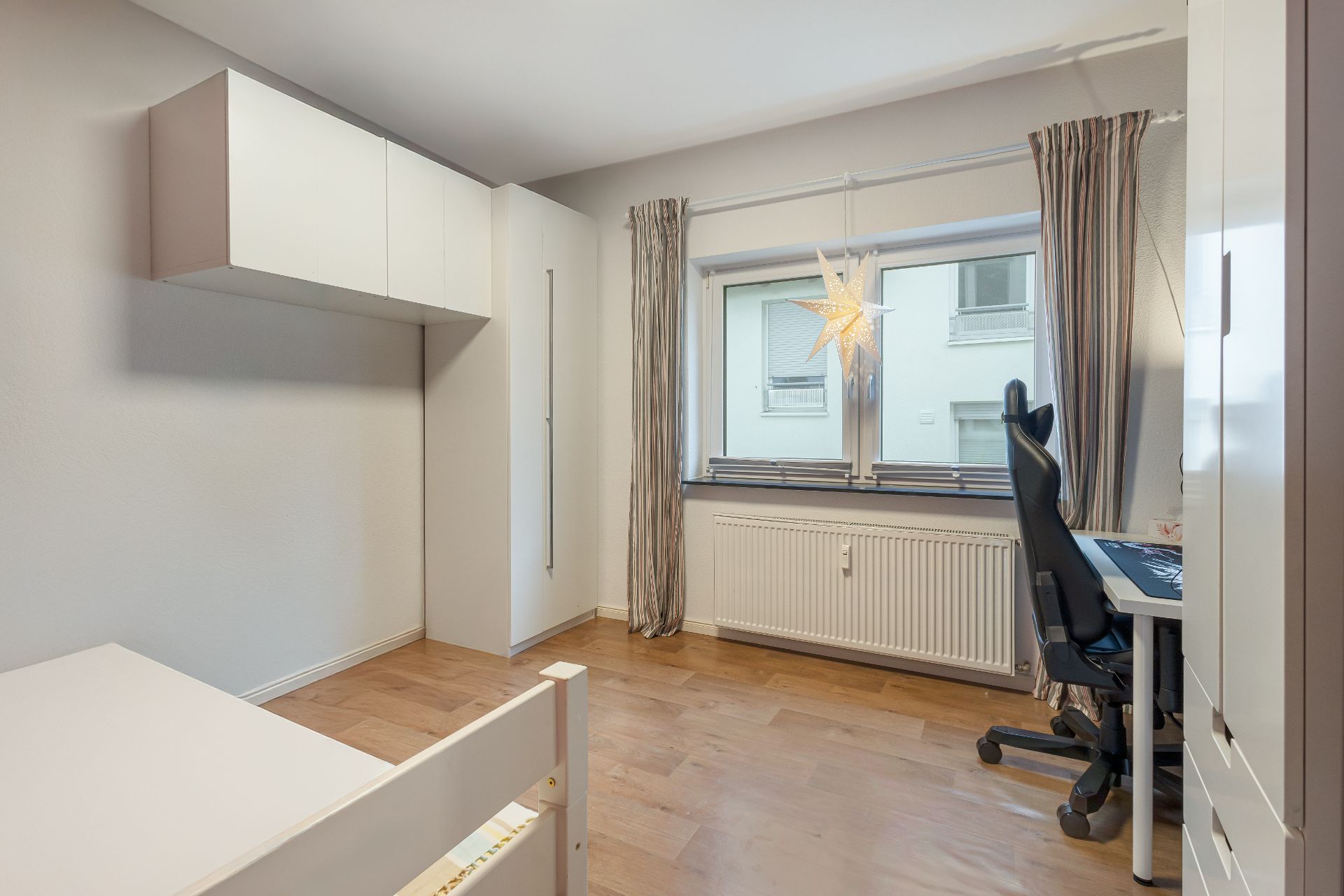 Helles Zimmer mit Holzboden, Schreibtisch, Stuhl, Fenster mit Vorhängen und Hängeschrank.