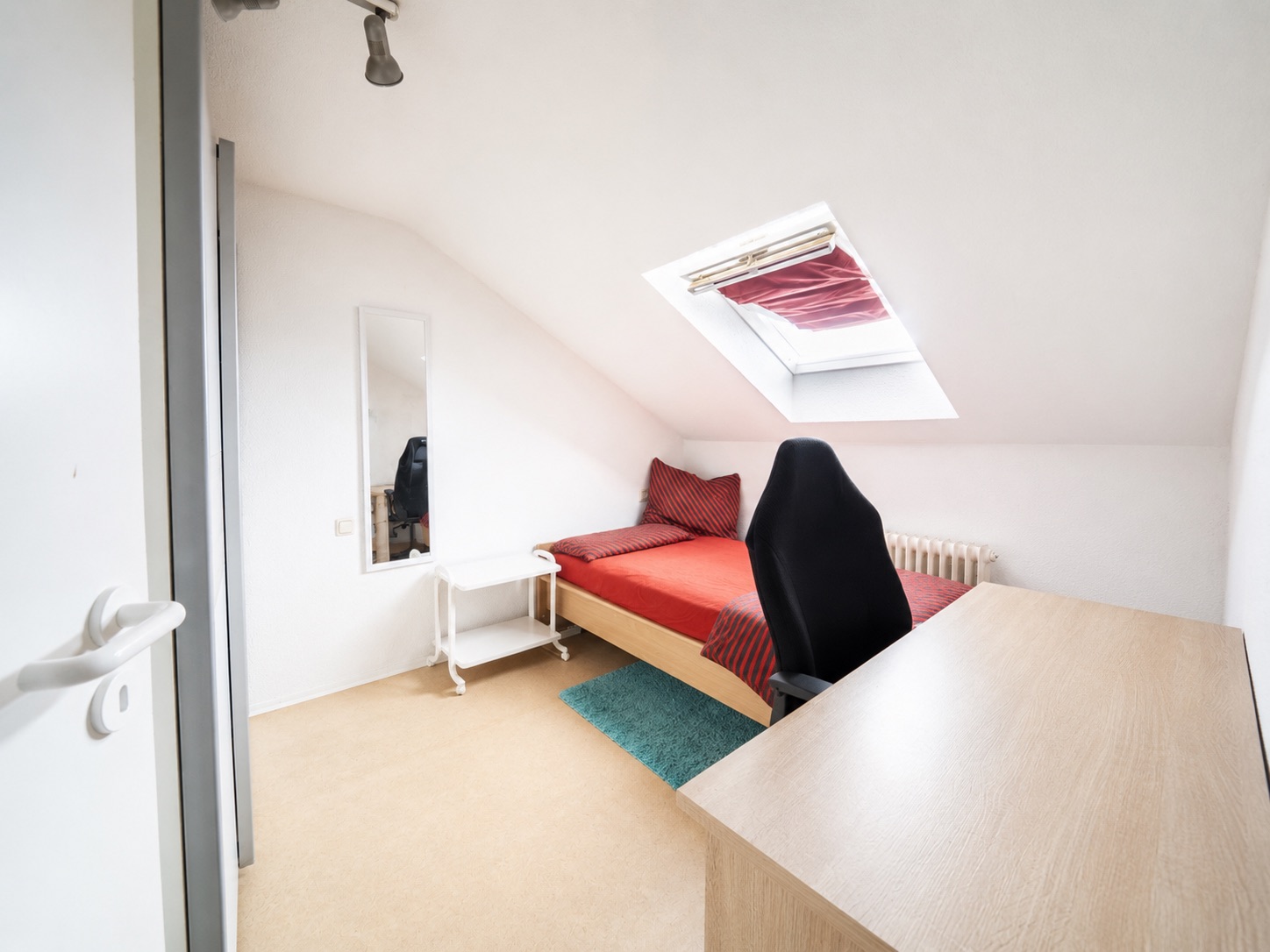Kleines Zimmer mit Schreibtisch, Einzelbett, Dachfenster und Spiegel an der Wand.