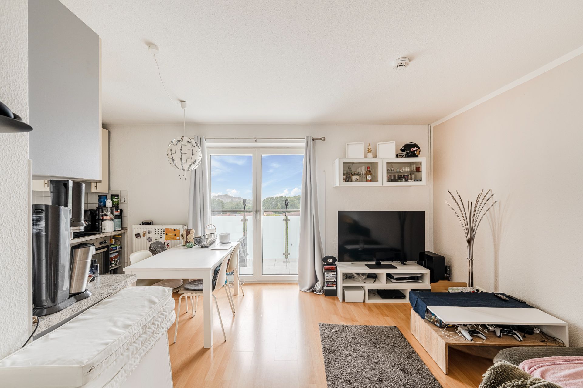 Helles Wohnzimmer mit Esstisch, Fernseher, Regal und Zugang zu einem Balkon.