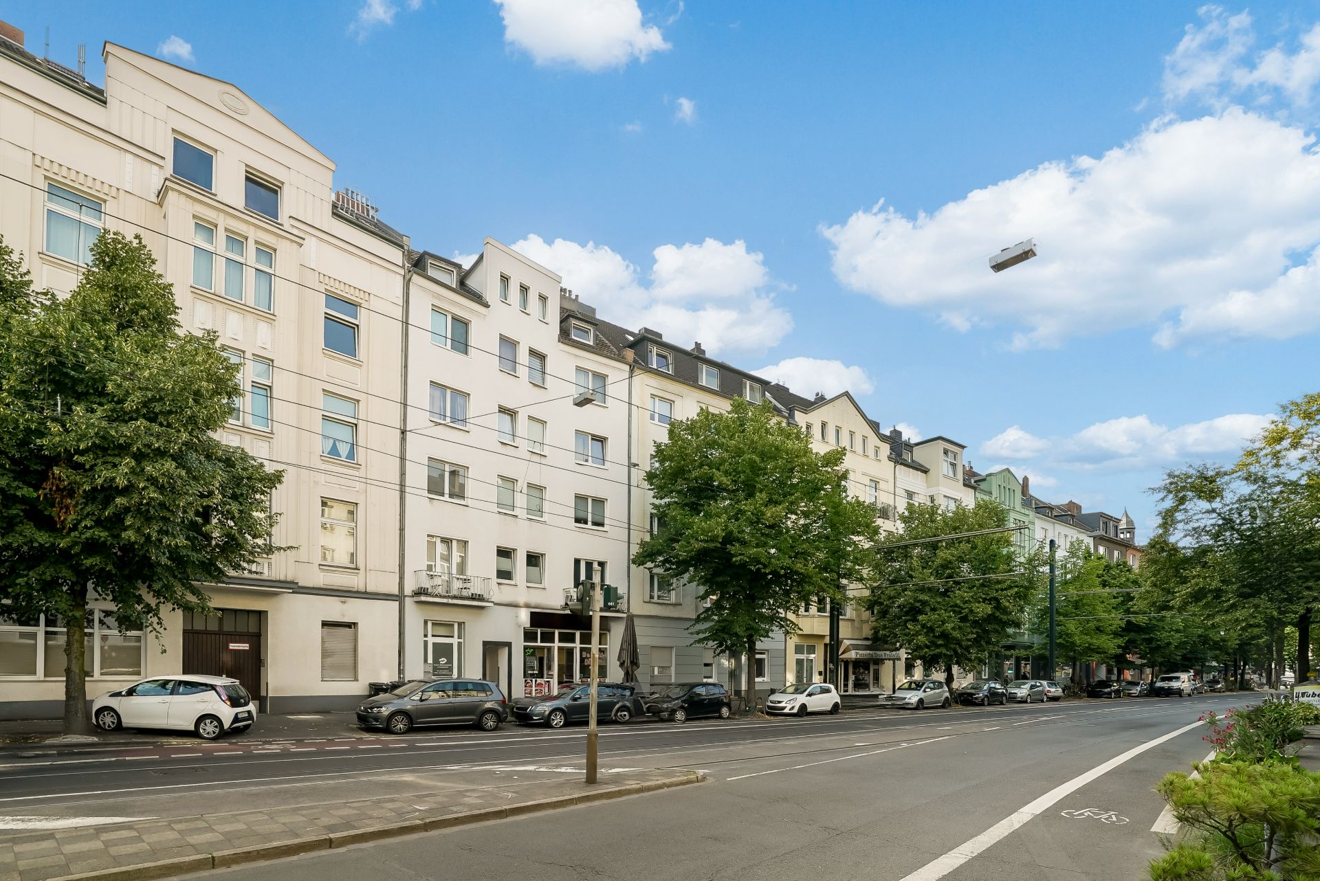 Mehrstöckige Wohnhäuser an einer Straße mit Autos und Bäumen, blauer Himmel mit Wolken.