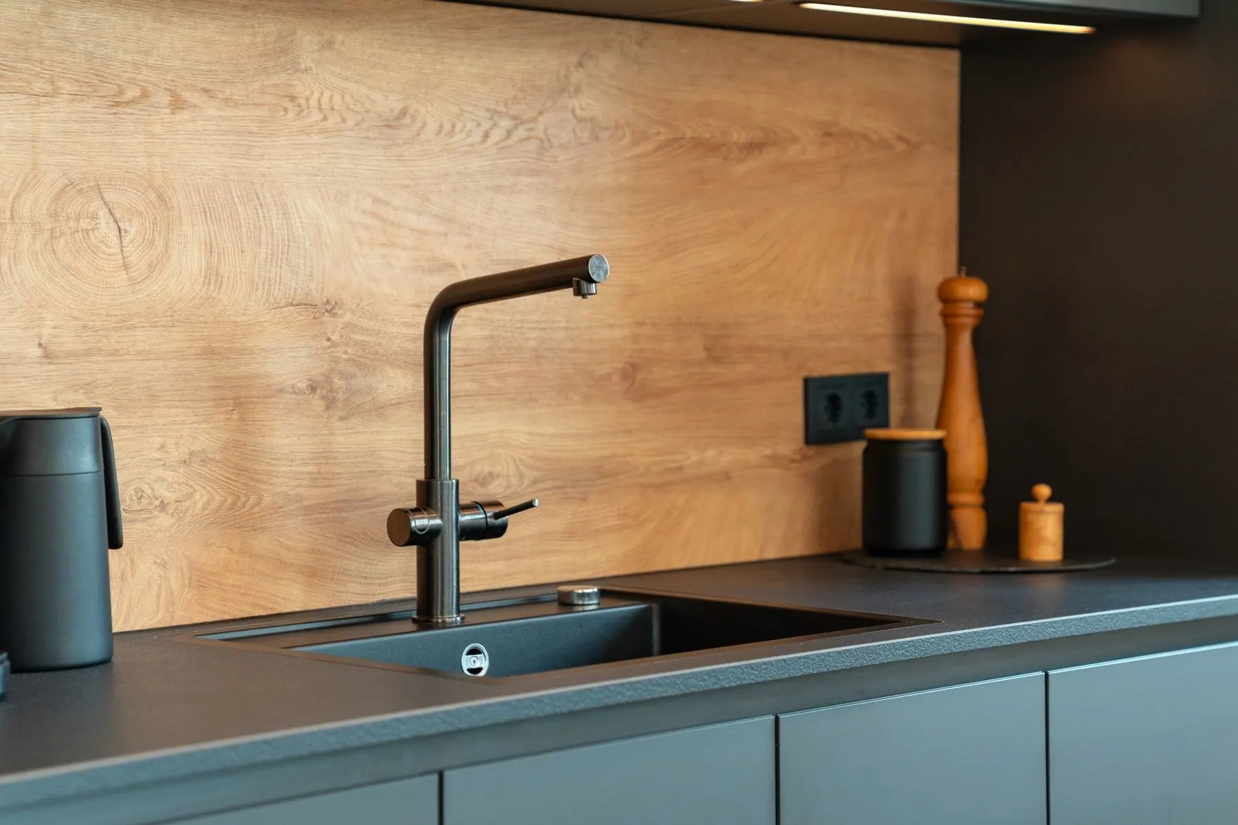 Moderne Küchenspüle mit schwarzem Wasserhahn und Holzwand, daneben Küchenutensilien.