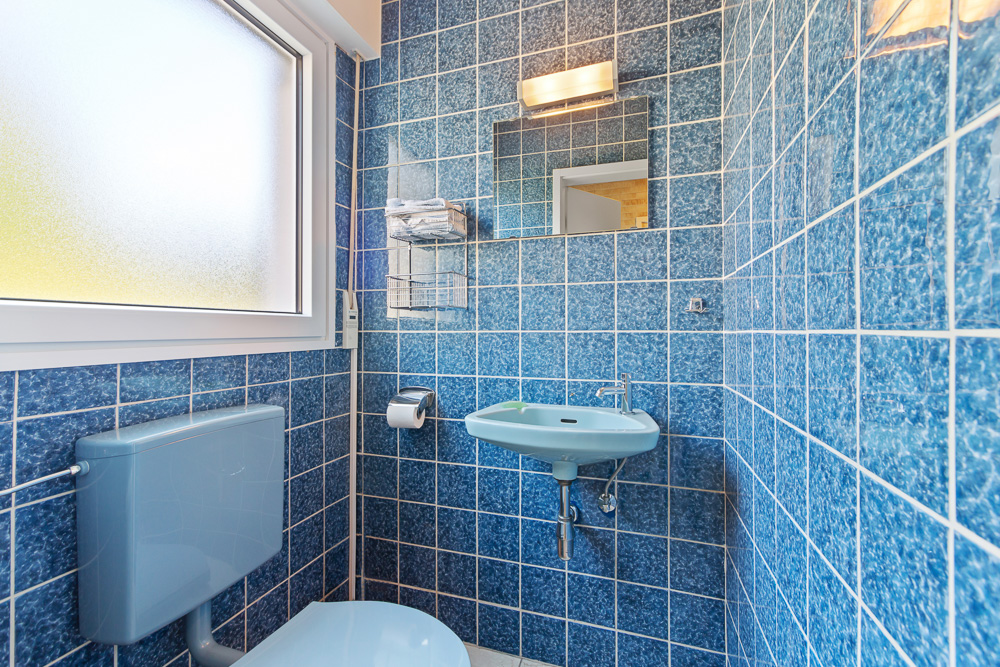 Kleines Badezimmer mit blauen Fliesen, Toilette, Waschbecken, Spiegel und Fenster.