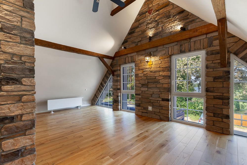 Helles Zimmer mit Holzfußboden, sichtbaren Holzbalken und Steinwand mit Fenstern.