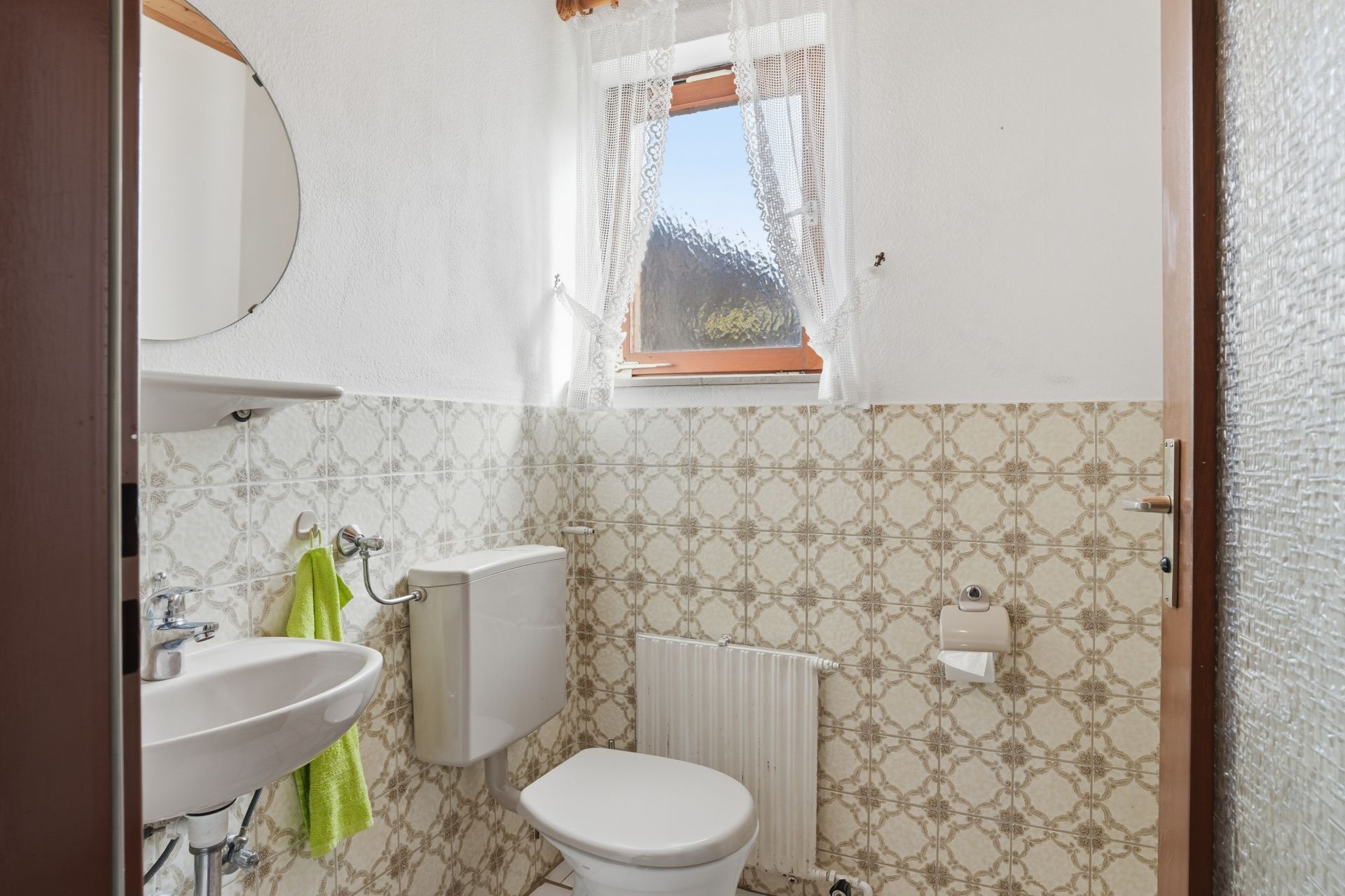 Kleines Badezimmer mit Toilette, Waschbecken, Fenster und gemusterten Fliesen an der Wand.