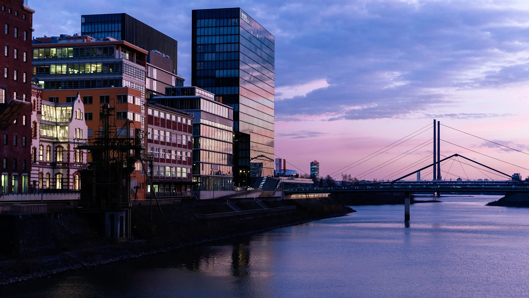 Moderne und historische Gebäude am Flussufer bei Sonnenuntergang, im Hintergrund eine Brücke.