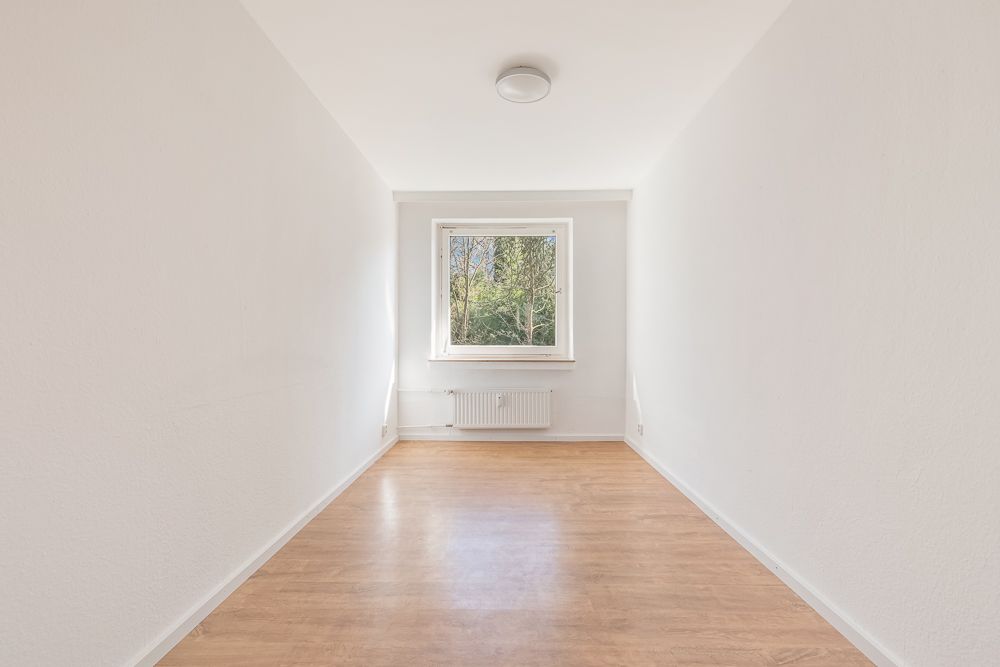 Heller, schmaler Raum mit Holzfußboden, weiß gestrichenen Wänden und einem Fenster am Ende.