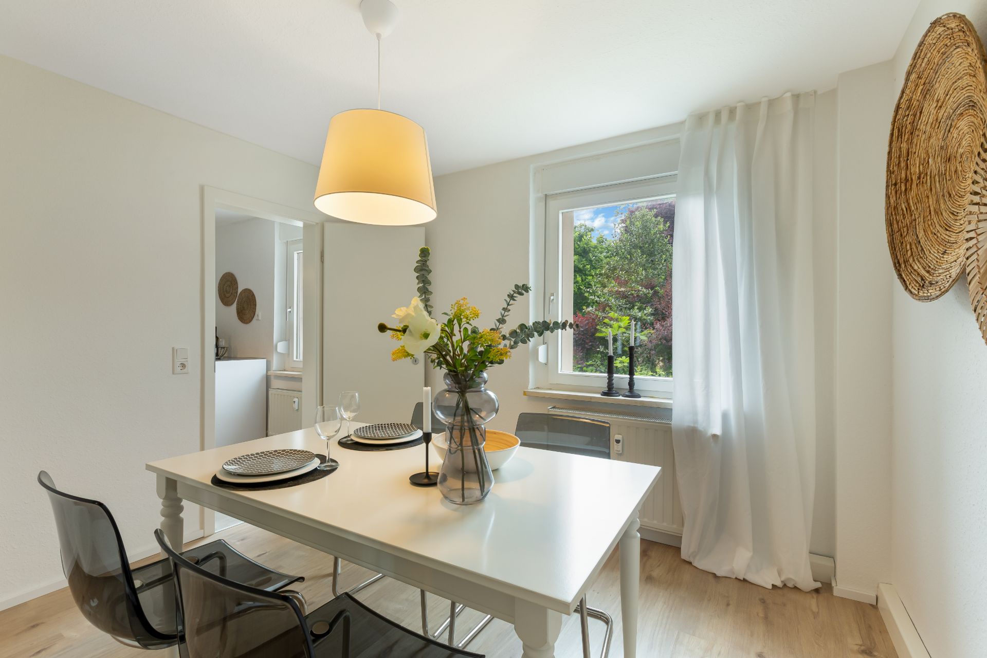 Heller Essbereich mit weißem Tisch, zwei Stühlen, Blumen und Fenster mit Vorhang.