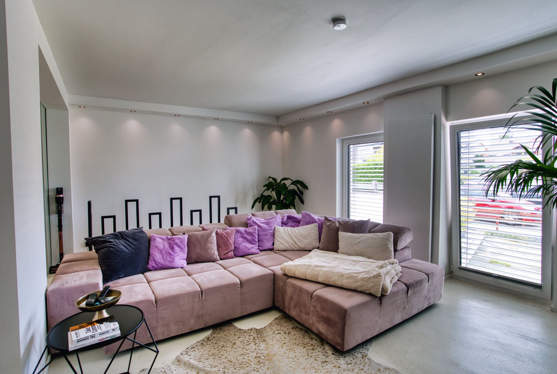 Modernes Wohnzimmer mit großer Eckcouch, vielen Kissen, Teppich und Pflanzen, große Fenster im Hintergrund.