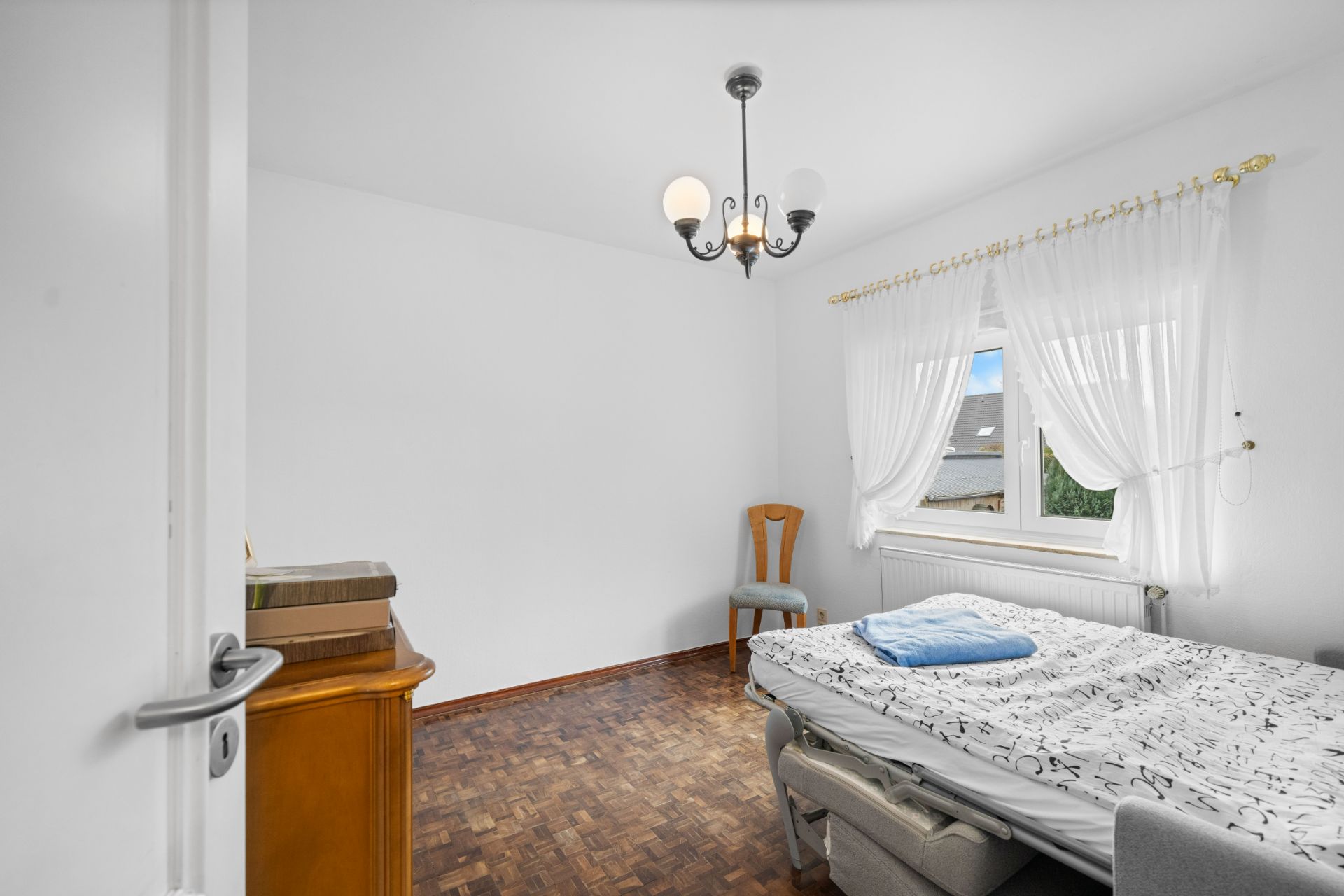 Helles Schlafzimmer mit Bett, Stuhl am Fenster, Holzboden und Deckenlampe.