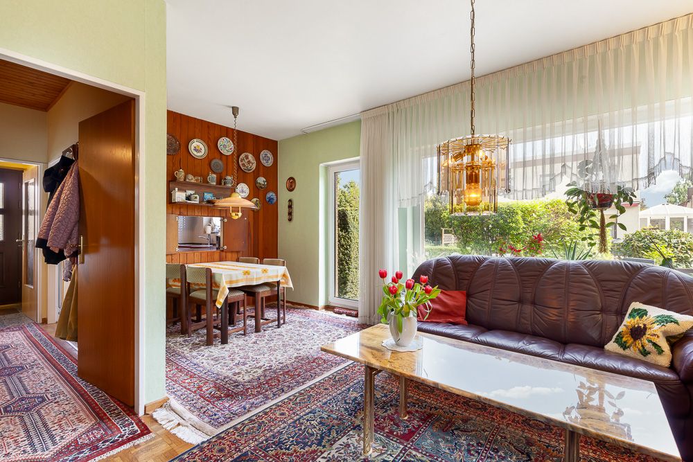 Wohnzimmer mit braunem Sofa, Couchtisch und Essbereich am Fenster, bunte Teppiche auf dem Boden.