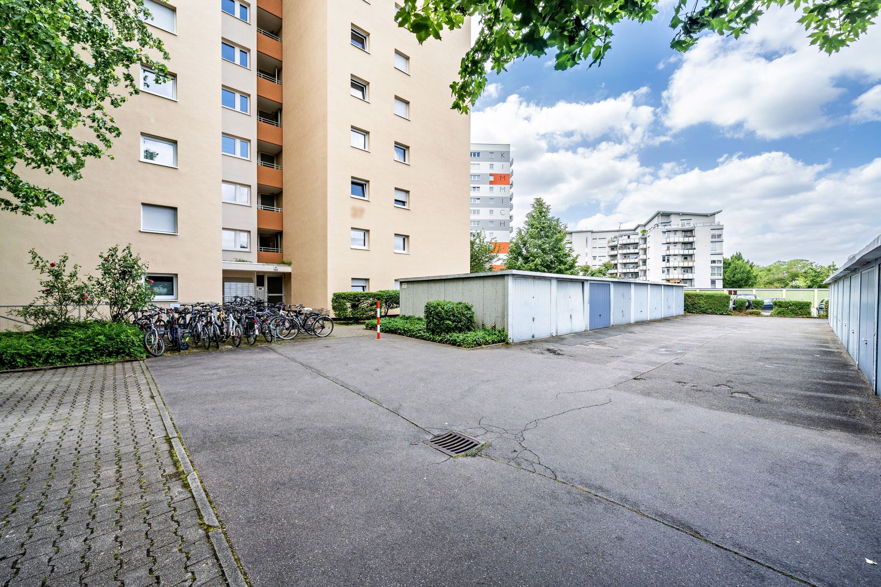 Mehrfamilienhaus mit Fahrradständern und Garagen auf einem asphaltierten Hof.
