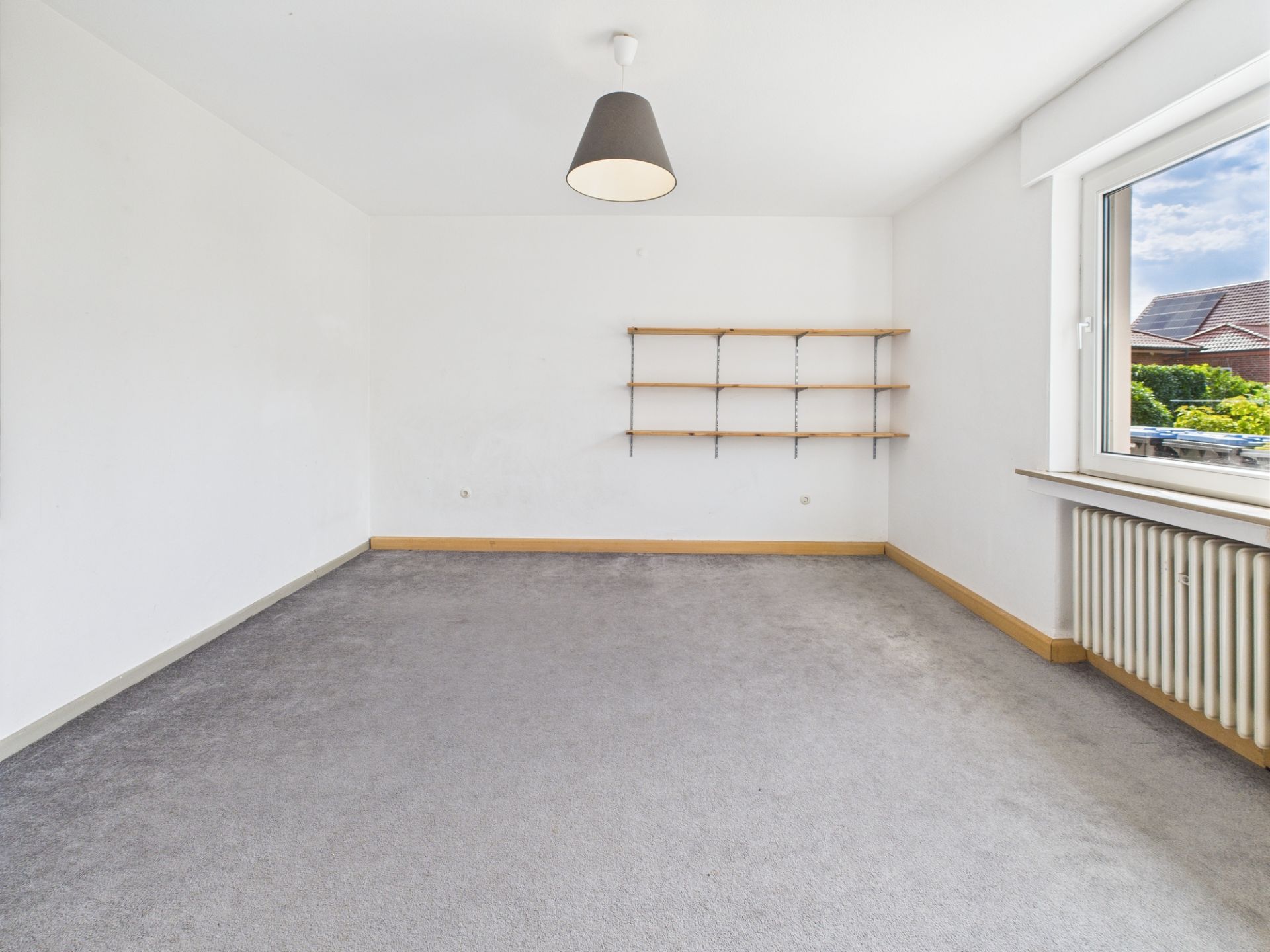 Leeres Zimmer mit grauem Teppich, Wandregalen und großem Fenster.