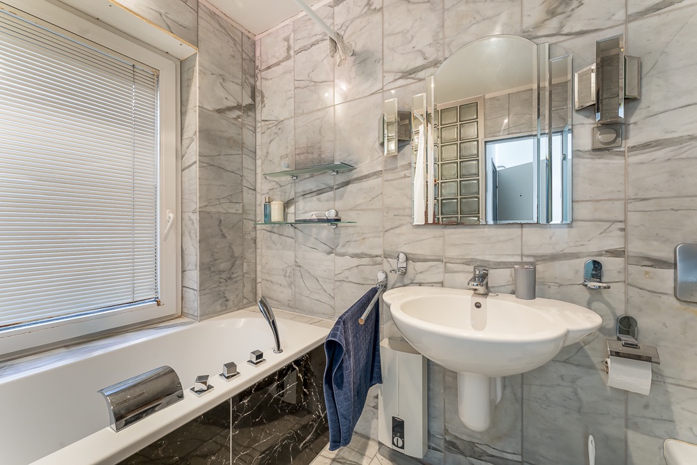 Modernes Bad mit Marmorfliesen, Badewanne, Waschbecken, Spiegel und Fenster mit Jalousie.