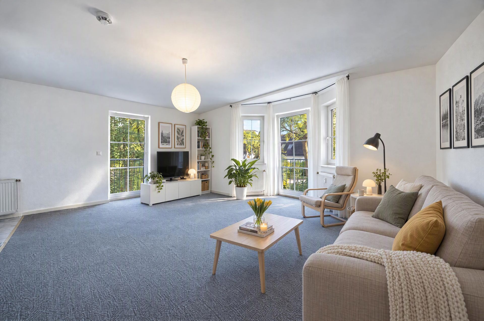 Helles Wohnzimmer mit Sofa, Couchtisch, Sessel, TV und großen Fenstern mit Blick ins Grüne.