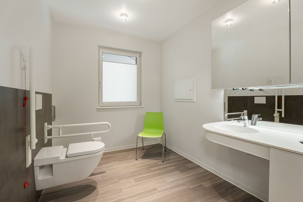 Barrierefreies Badezimmer mit Haltegriffen, grünem Stuhl, Toilette und Waschbecken.