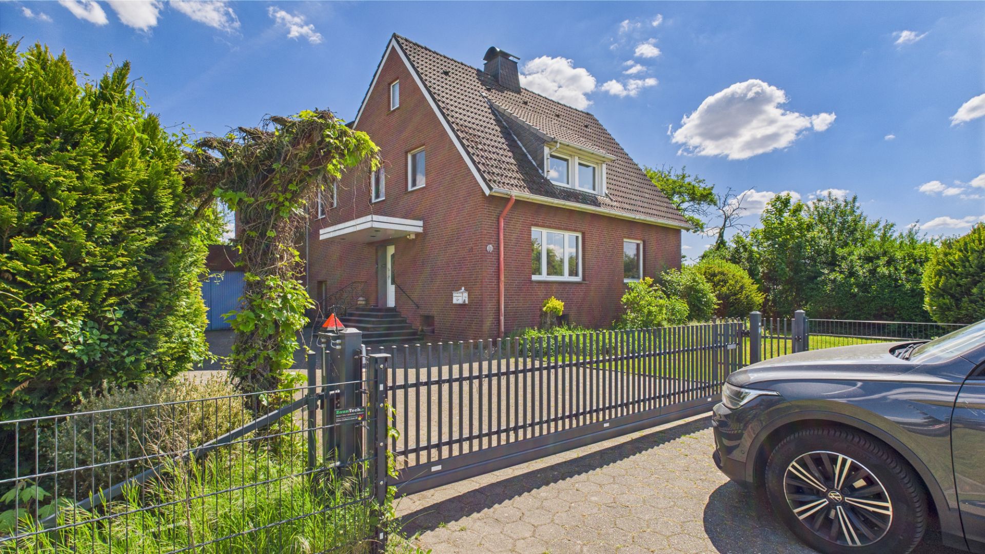 Zweistöckiges rotes Backsteinhaus mit Garten, Einfahrtstor und geparktem Auto vorne.