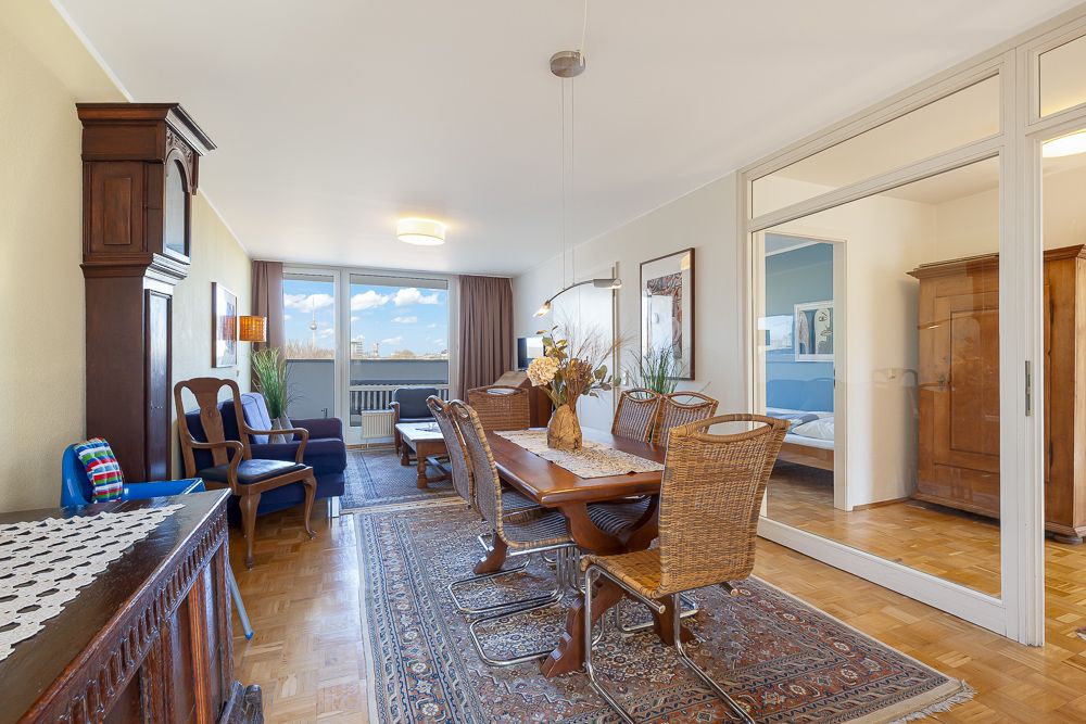 Helles Wohnzimmer mit Holzmöbeln, Esstisch, Teppich und Zugang zu einem Schlafzimmer.