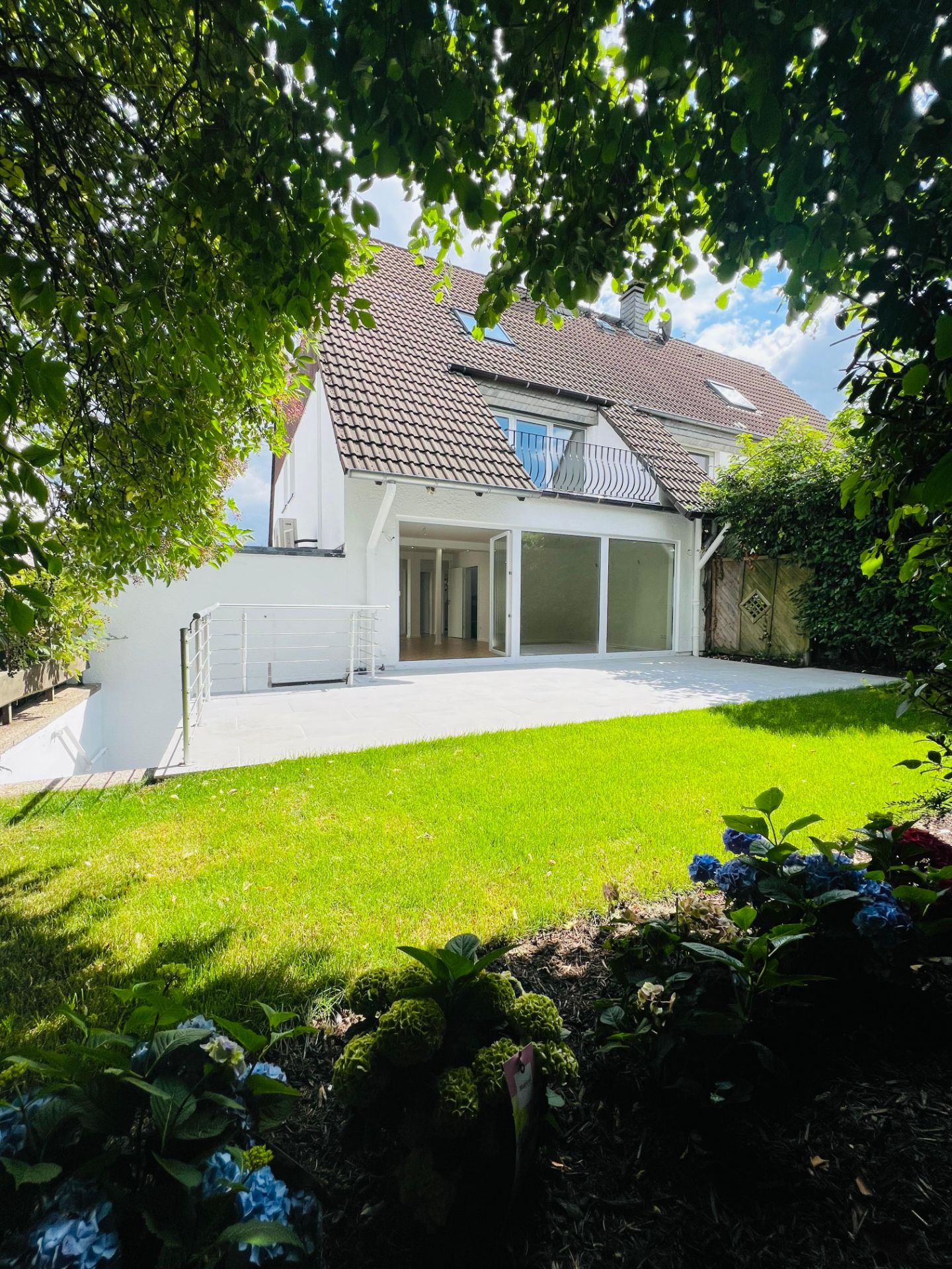 Blick auf ein Wohnhaus mit Terrasse und grünem Garten, umrahmt von Bäumen und Blumen.