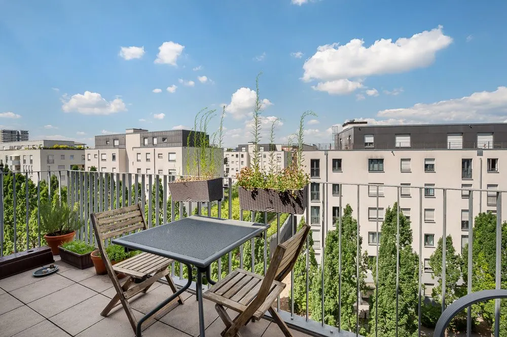 Balkon mit Holztisch, zwei Stühlen und Pflanzen, Blick auf umliegende Wohngebäude.