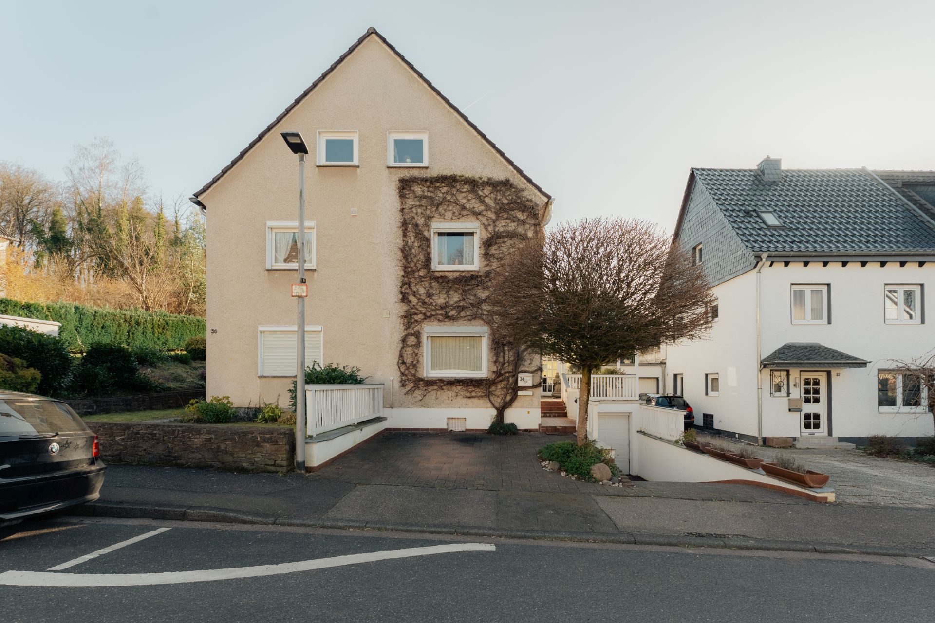 Beiges Wohnhaus mit Kletterpflanzen an der Fassade und Auffahrt, daneben ein weißes Haus.