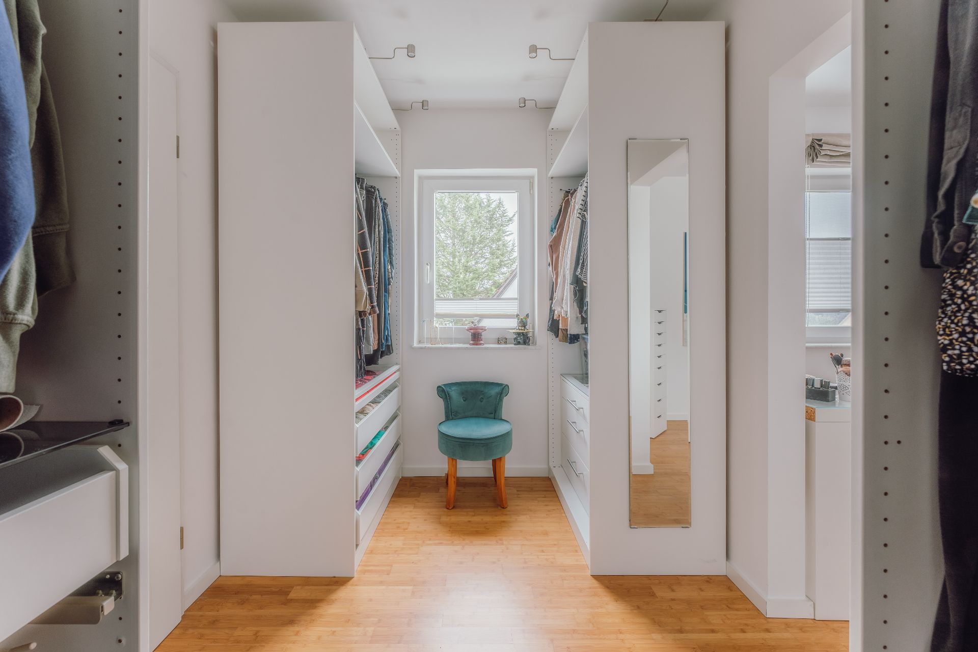 Heller begehbarer Kleiderschrank mit grünem Stuhl, Fenster und Spiegel an der Wand.