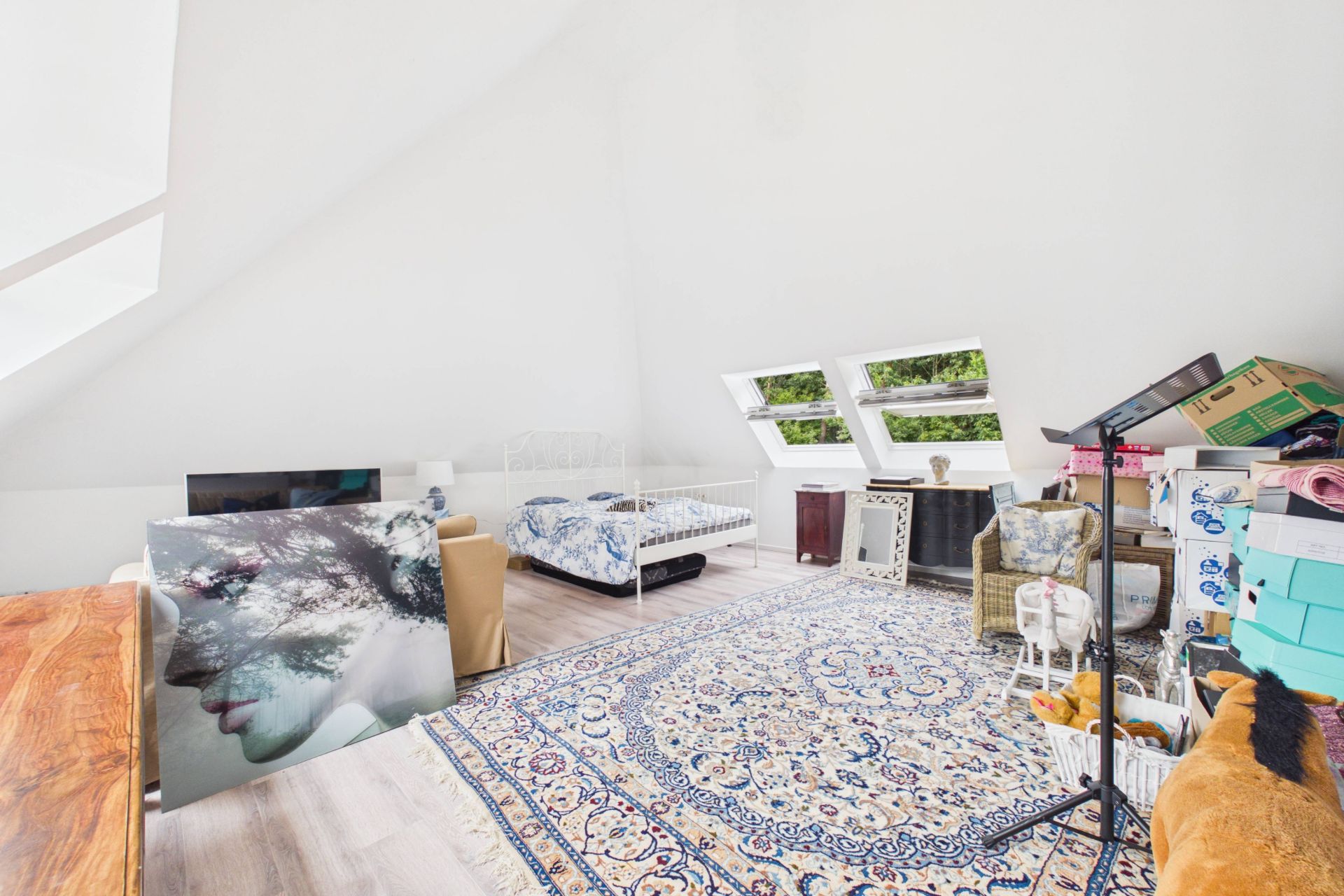 Helles Dachzimmer mit Bett, Teppich, Sesseln und mehreren Fenstern, dekoriert mit Bildern und Kisten.
