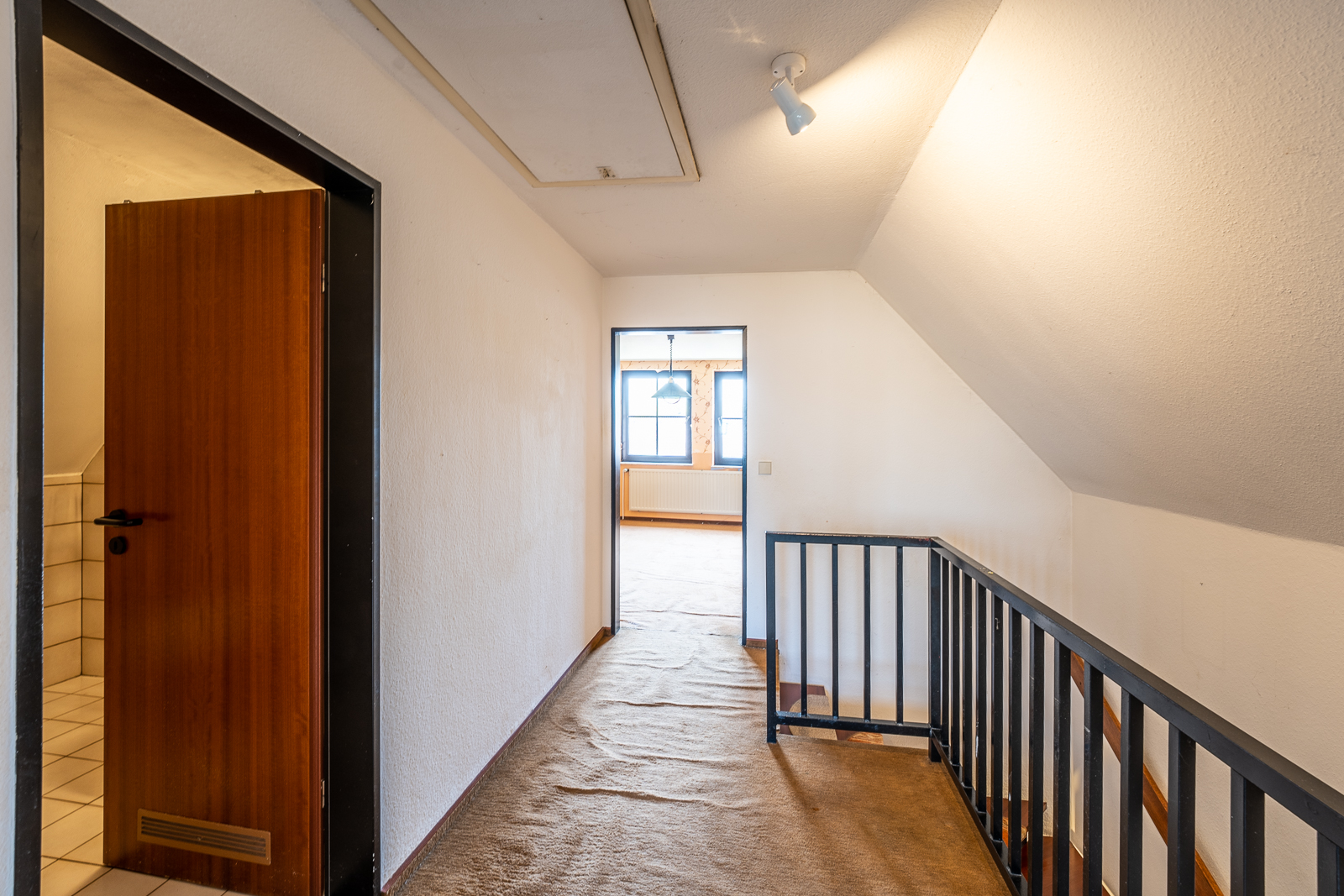 Flur mit Teppichboden, offener Tür zu einem Badezimmer und Treppengeländer, Fenster im Hintergrund.