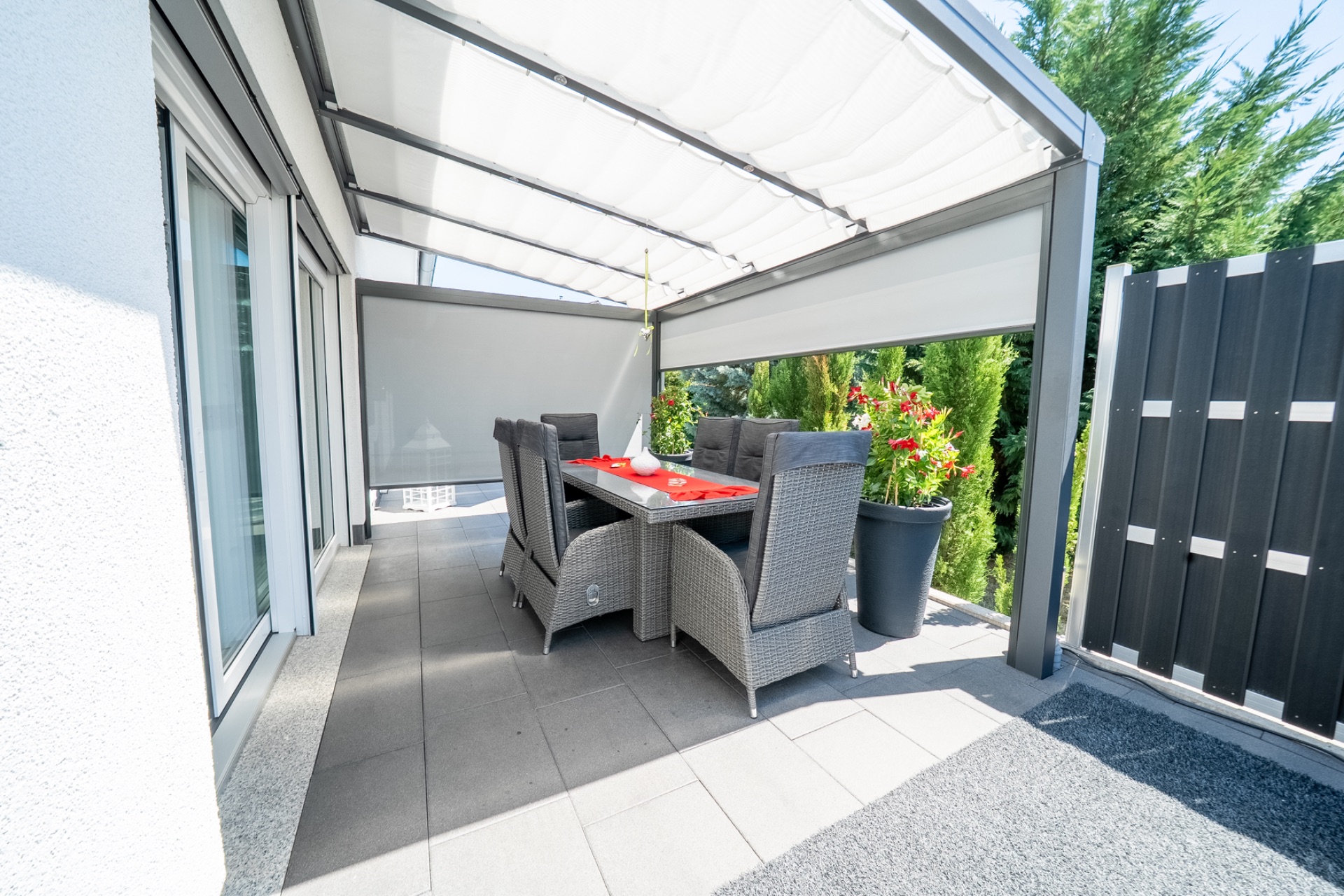 Überdachte Terrasse mit Esstisch und sechs Stühlen, umgeben von Pflanzen und Sichtschutz.