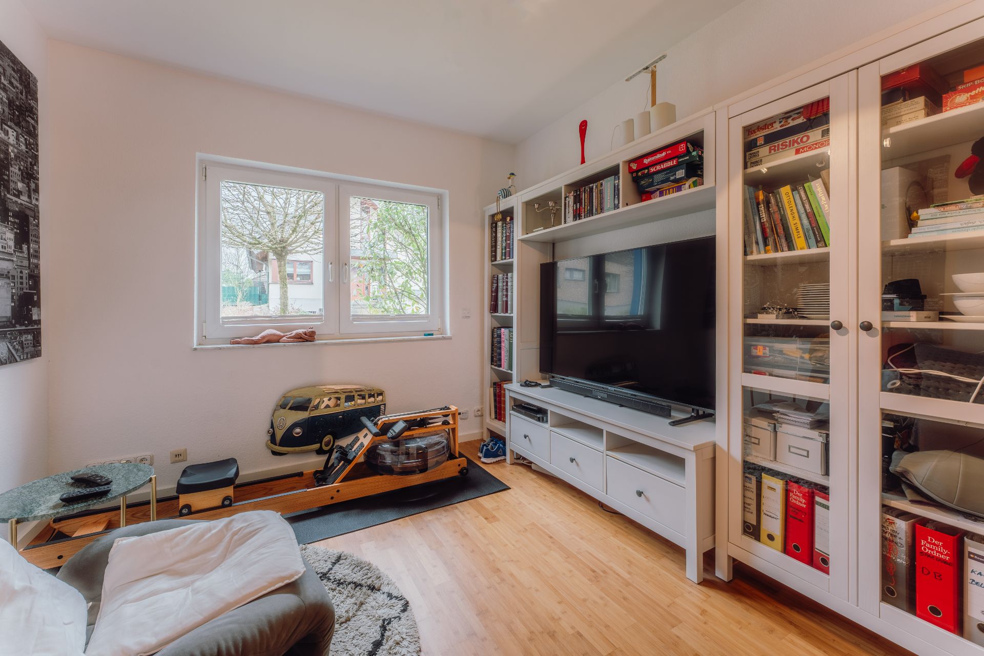 Wohnzimmer mit Fernseher, Bücherregalen, Sessel und Heimtrainer vor einem Fenster.