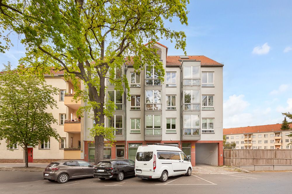 Mehrfamilienhaus mit vier Etagen, Bäumen davor und geparkten Autos auf der Straße.