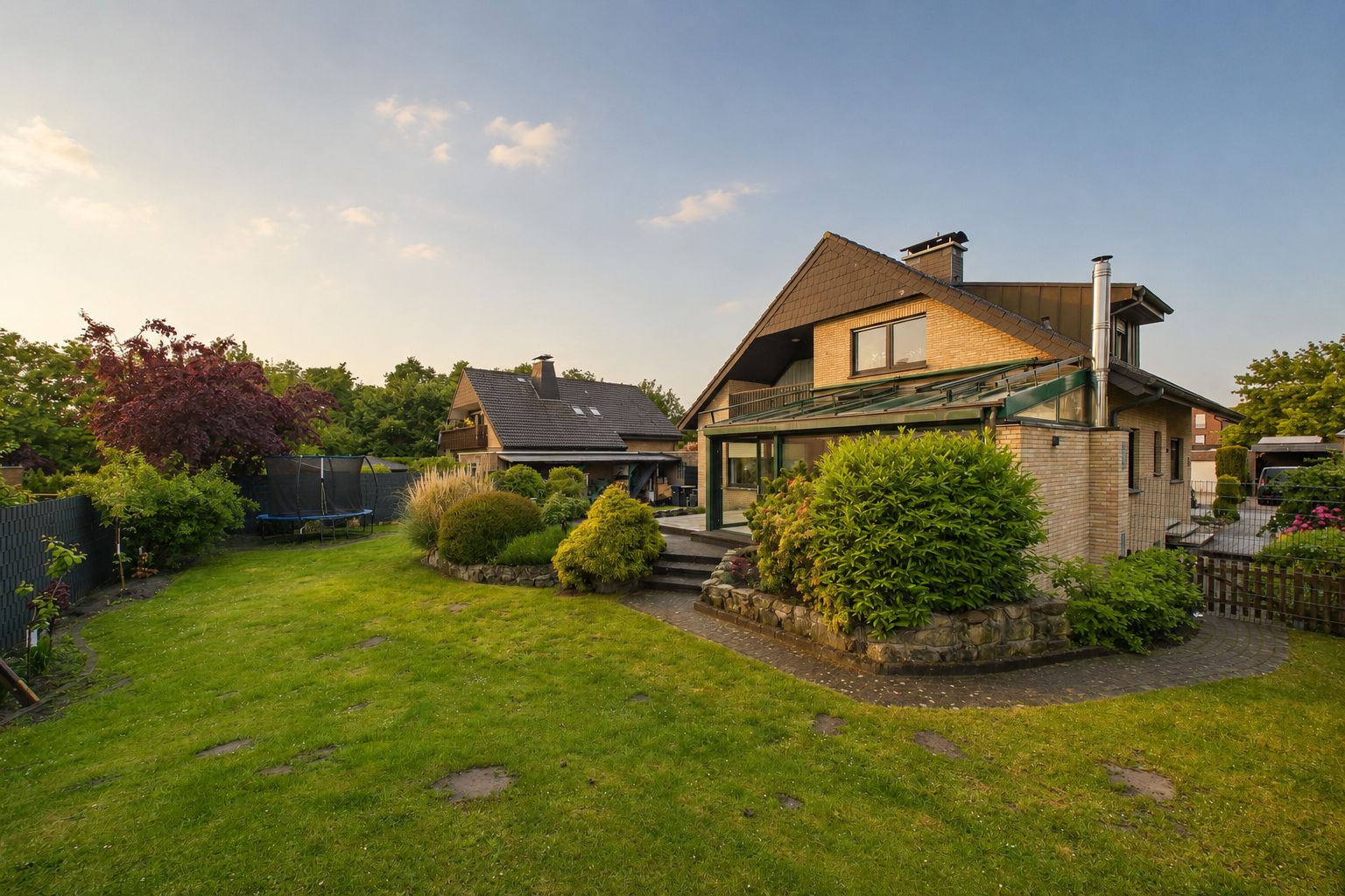 Einfamilienhaus mit großem Garten, Terrasse und verschiedenen Sträuchern bei Sonnenuntergang.