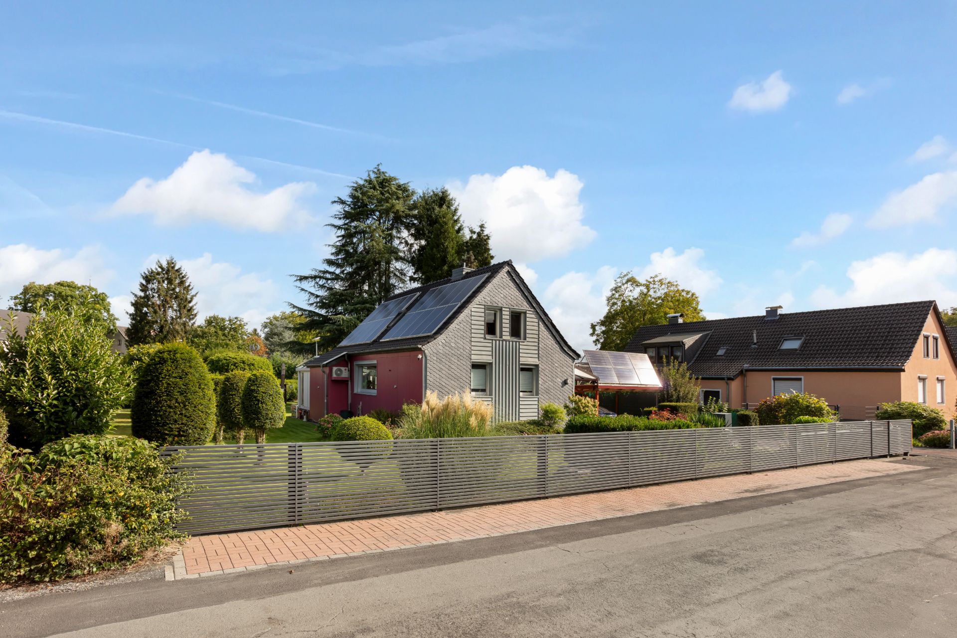 Modernes Haus mit Solarpanels, Garten und Metallzaun an einer ruhigen Wohnstraße.