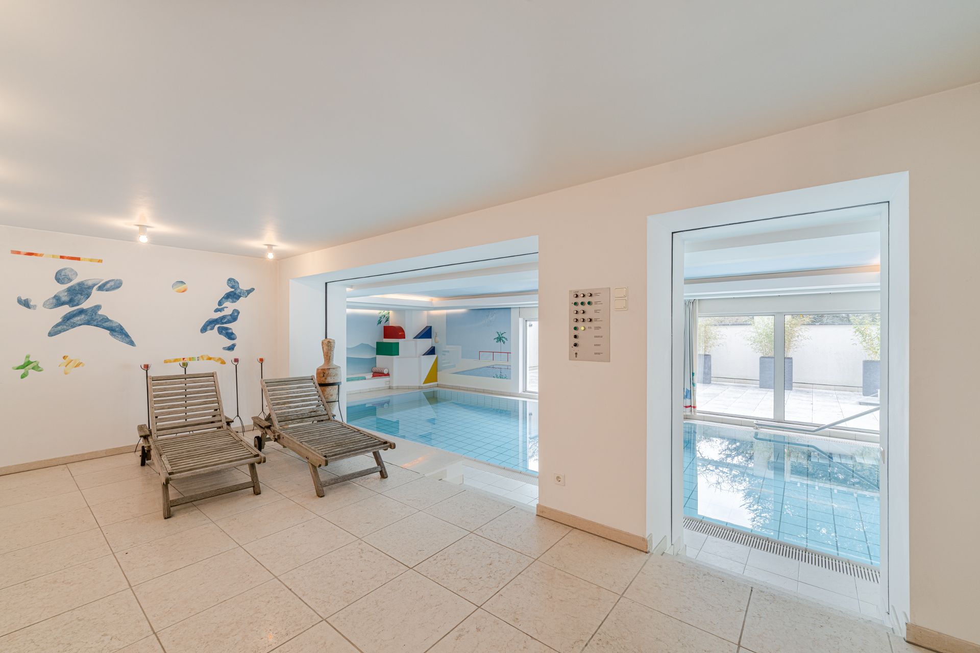 Innenraum mit zwei Liegestühlen und Blick auf einen Pool, Wand mit bunter Kunst.