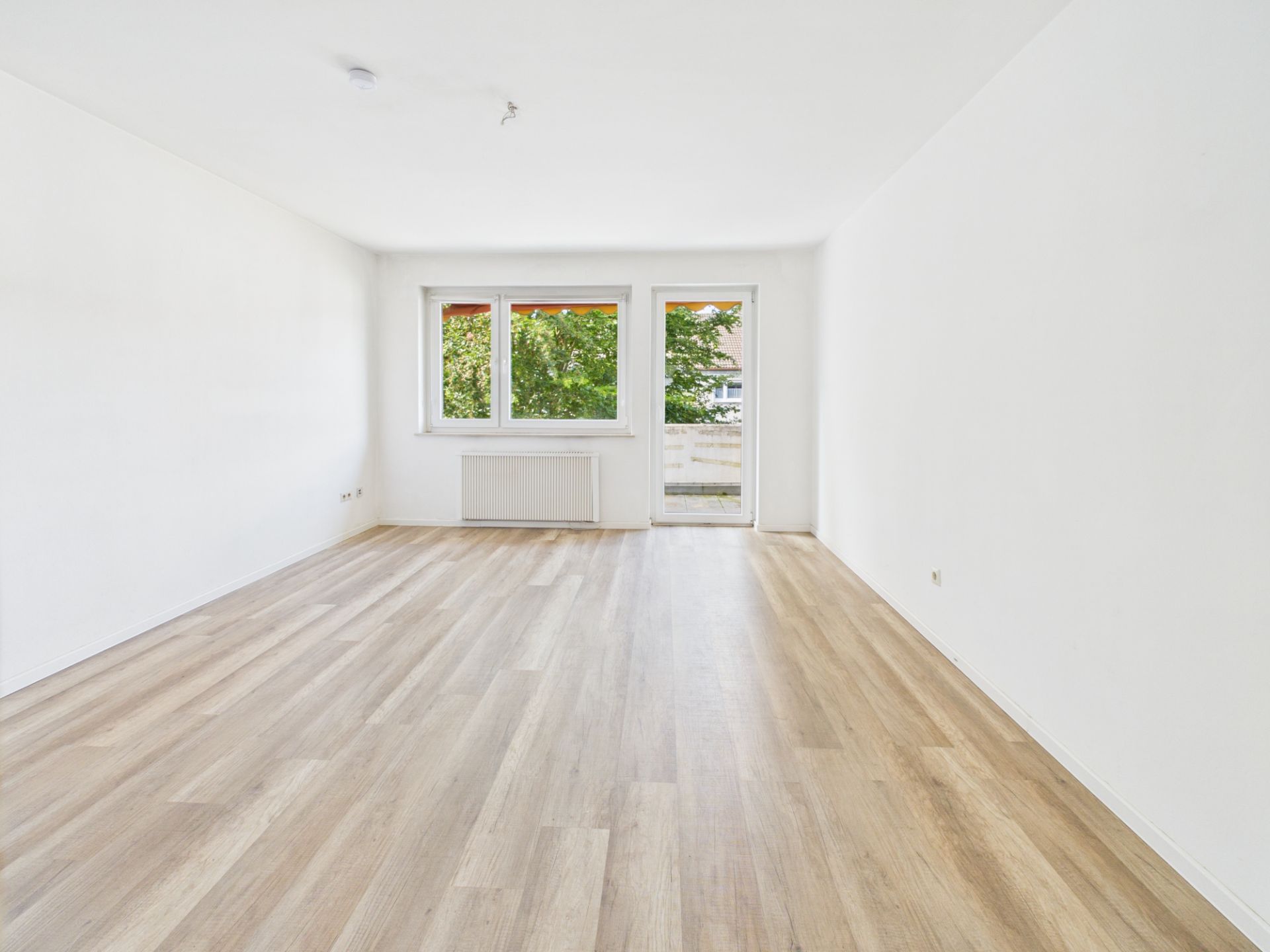 Helles, leeres Zimmer mit Holzfußboden, weißen Wänden, Fenstern und Tür zum Balkon.