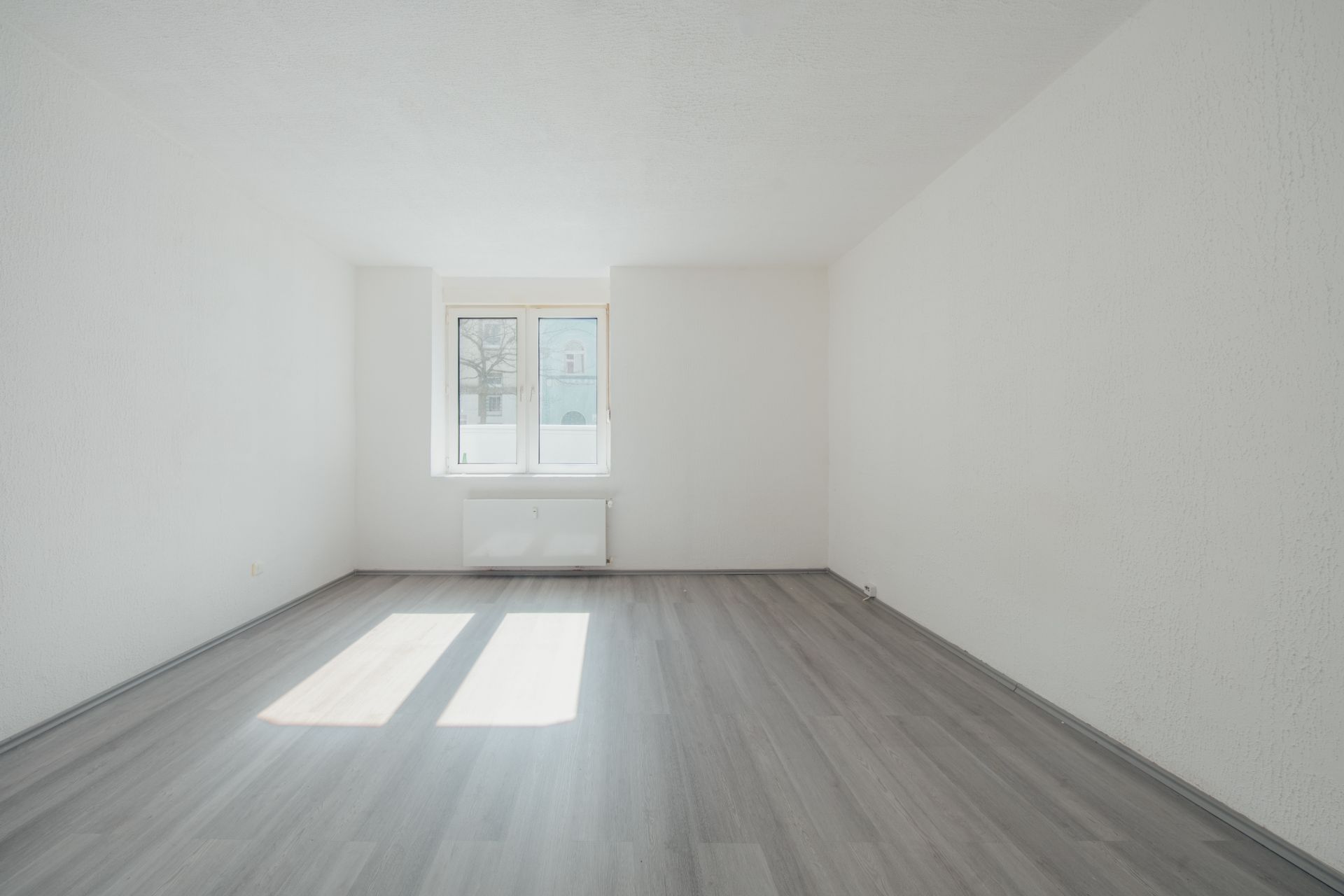 Leerer, heller Raum mit grauem Boden, weißen Wänden und einem Fenster an der Stirnseite.