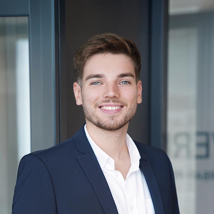 Ein junger Mann im Anzug steht lächelnd vor einer Glastür.