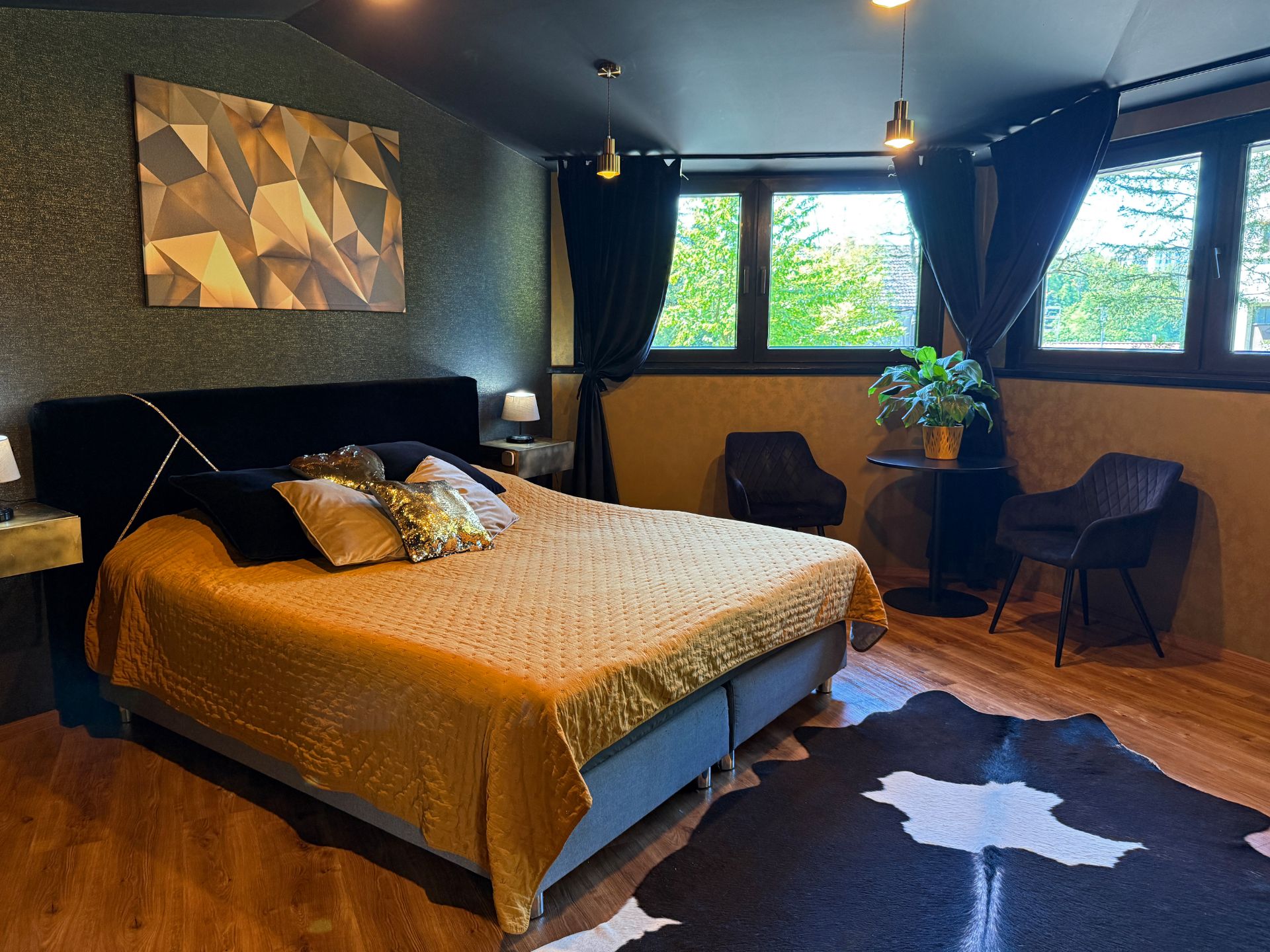 Modernes Schlafzimmer mit großem Bett, Wandkunst, zwei Sesseln, Lampe und großem Fenster mit Vorhängen.
