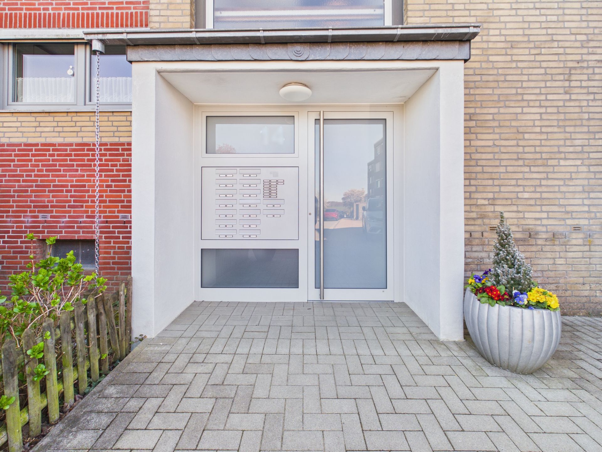 Eingangsbereich mit Briefkästen, Glastür und einem großen Blumenkübel mit bunten Blumen.