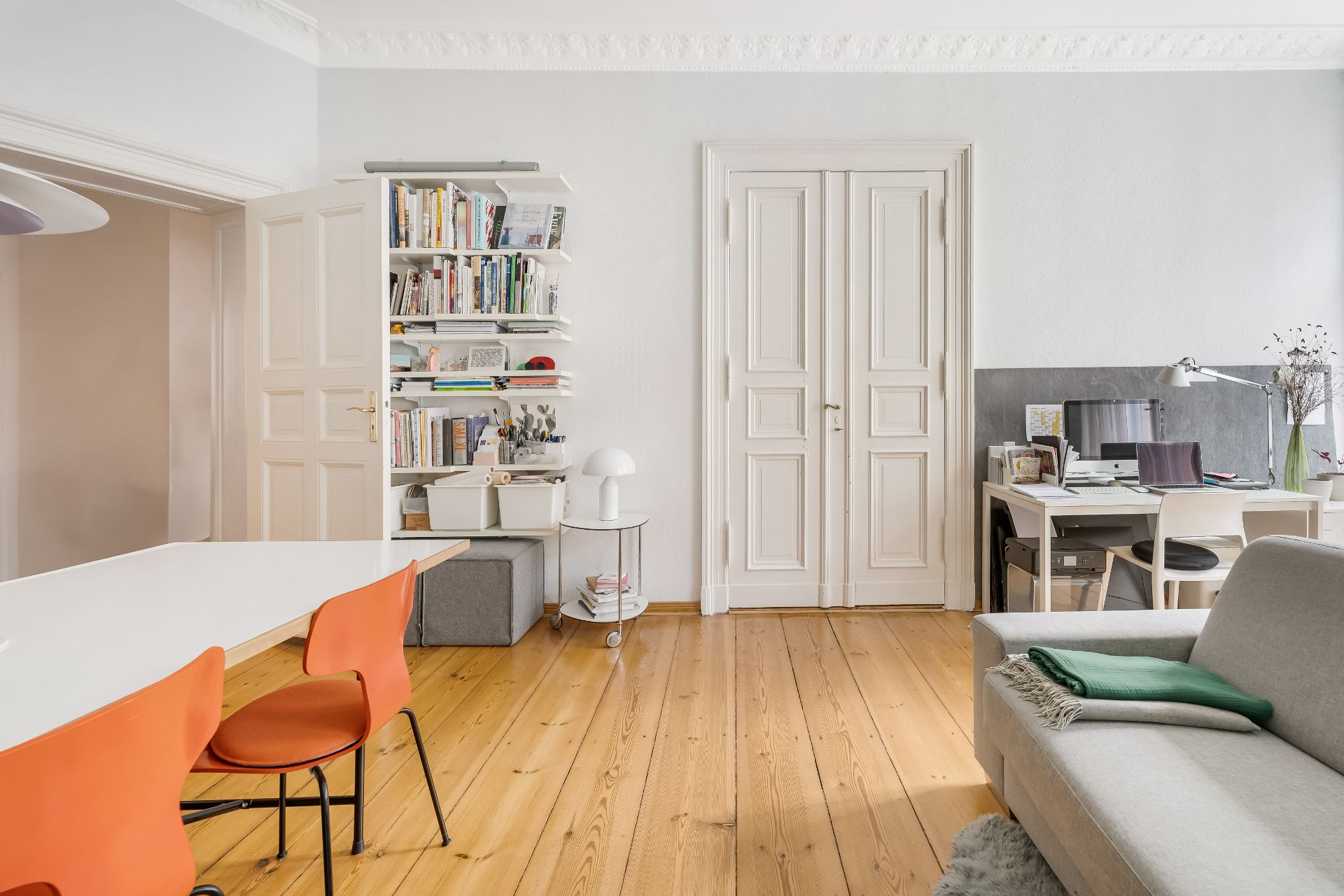 Helles Wohnzimmer mit Holzboden, Bücherregal, Schreibtisch, Sofa und Esstisch mit orangefarbenen Stühlen.