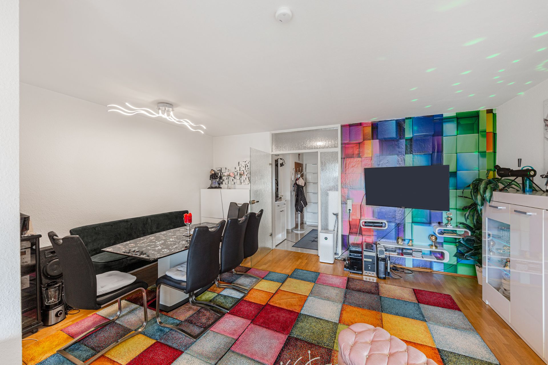 Wohnzimmer mit buntem Teppich, Esstisch, Stühlen und bunter Wand mit Fernseher.