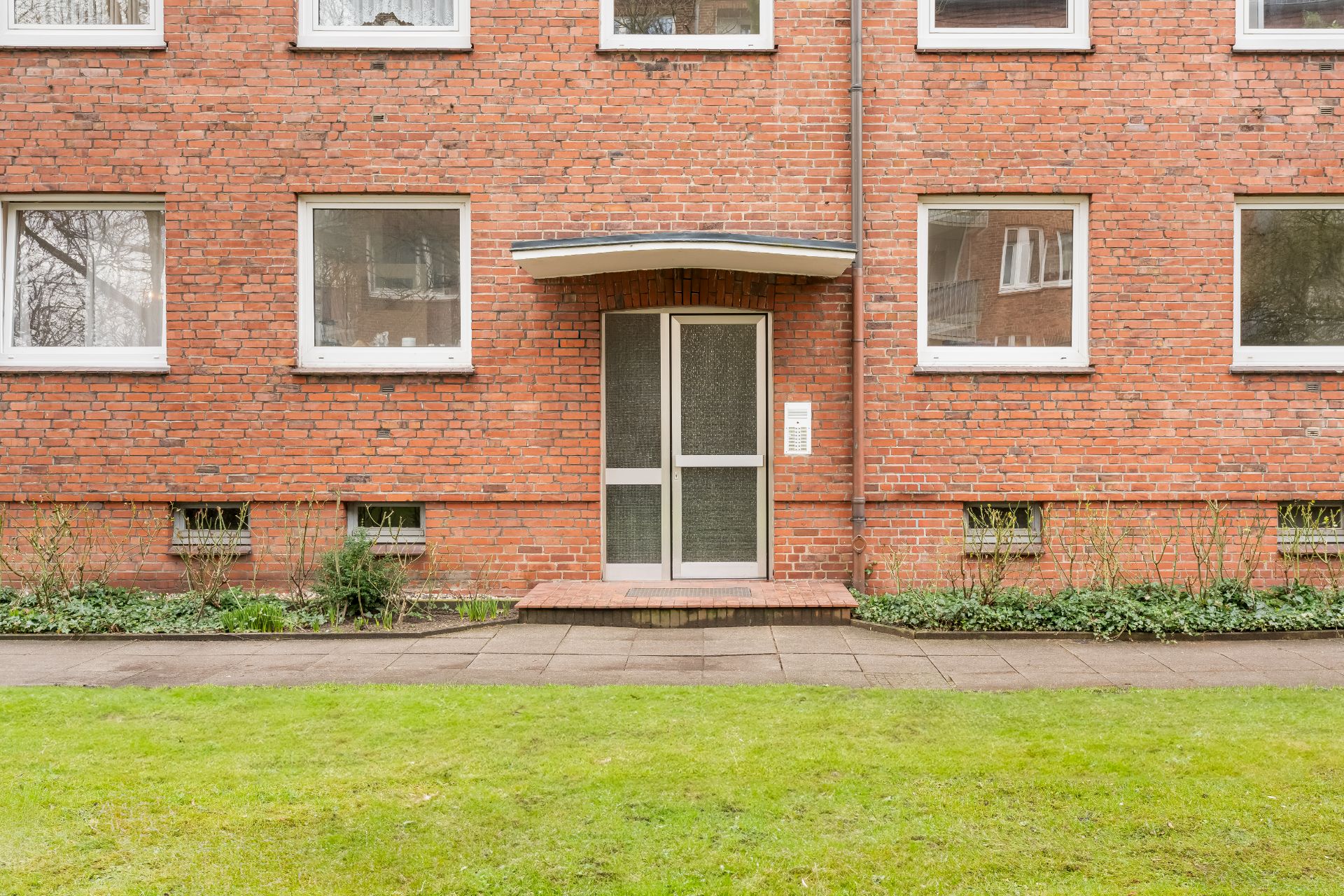 Eingangstür eines roten Mehrfamilienhauses mit Gehweg und Rasen davor.