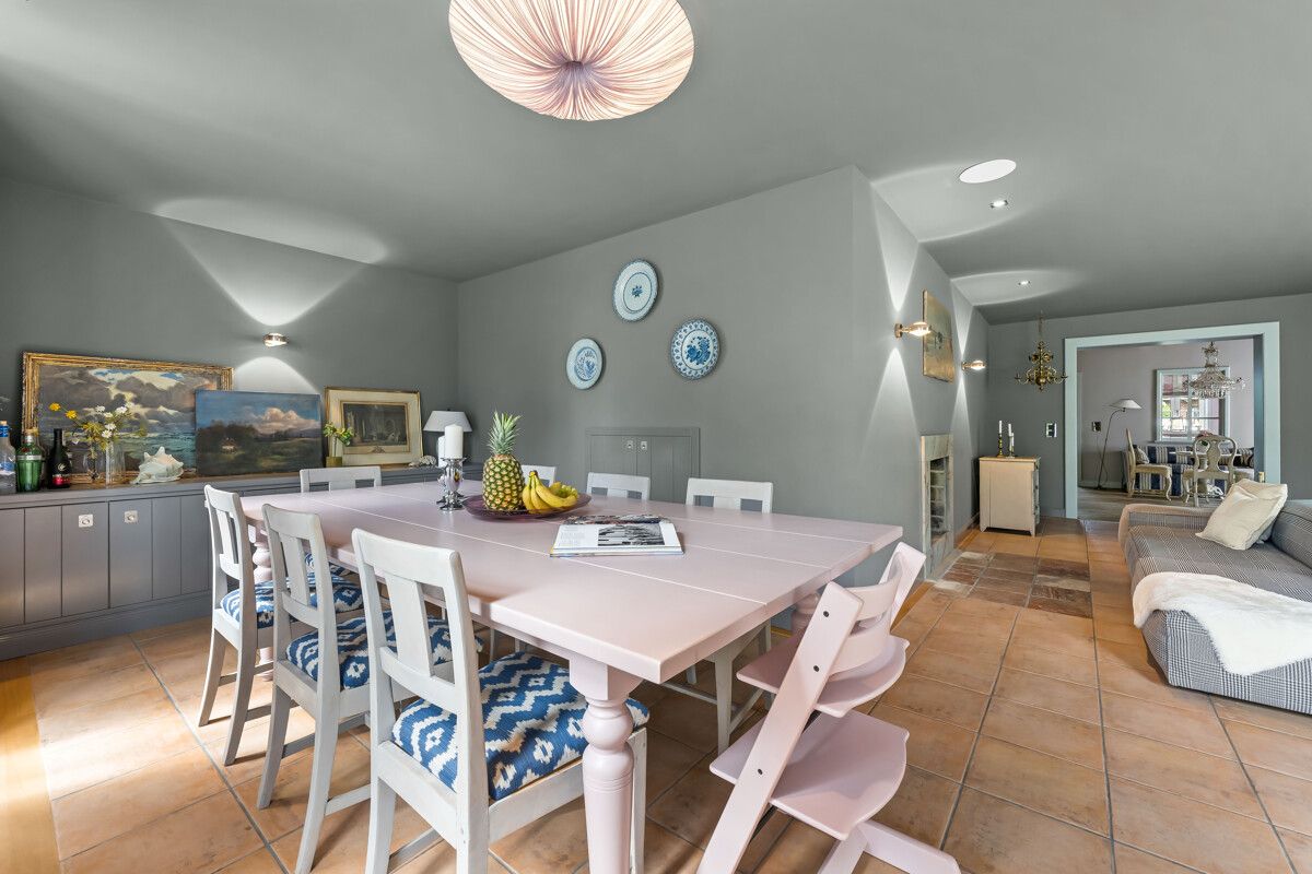 Esszimmer mit großem Tisch, weißen Stühlen und hellgrauen Wänden, im Hintergrund Wohnzimmer sichtbar.
