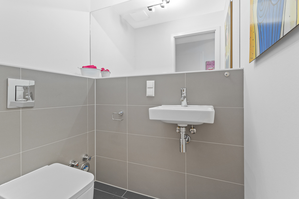 Modernes Gäste-WC mit grauen Fliesen, Wandtoilette, Waschbecken und Wandbild.