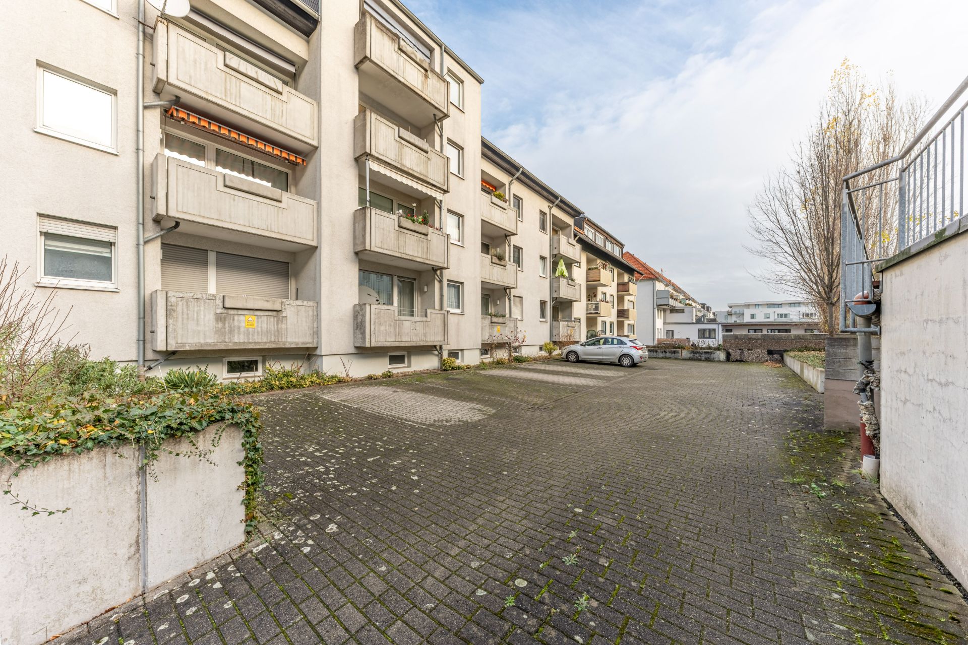 Gepflasterter Parkplatz vor einem mehrstöckigen Wohnhaus mit Balkonen und einem geparkten Auto.
