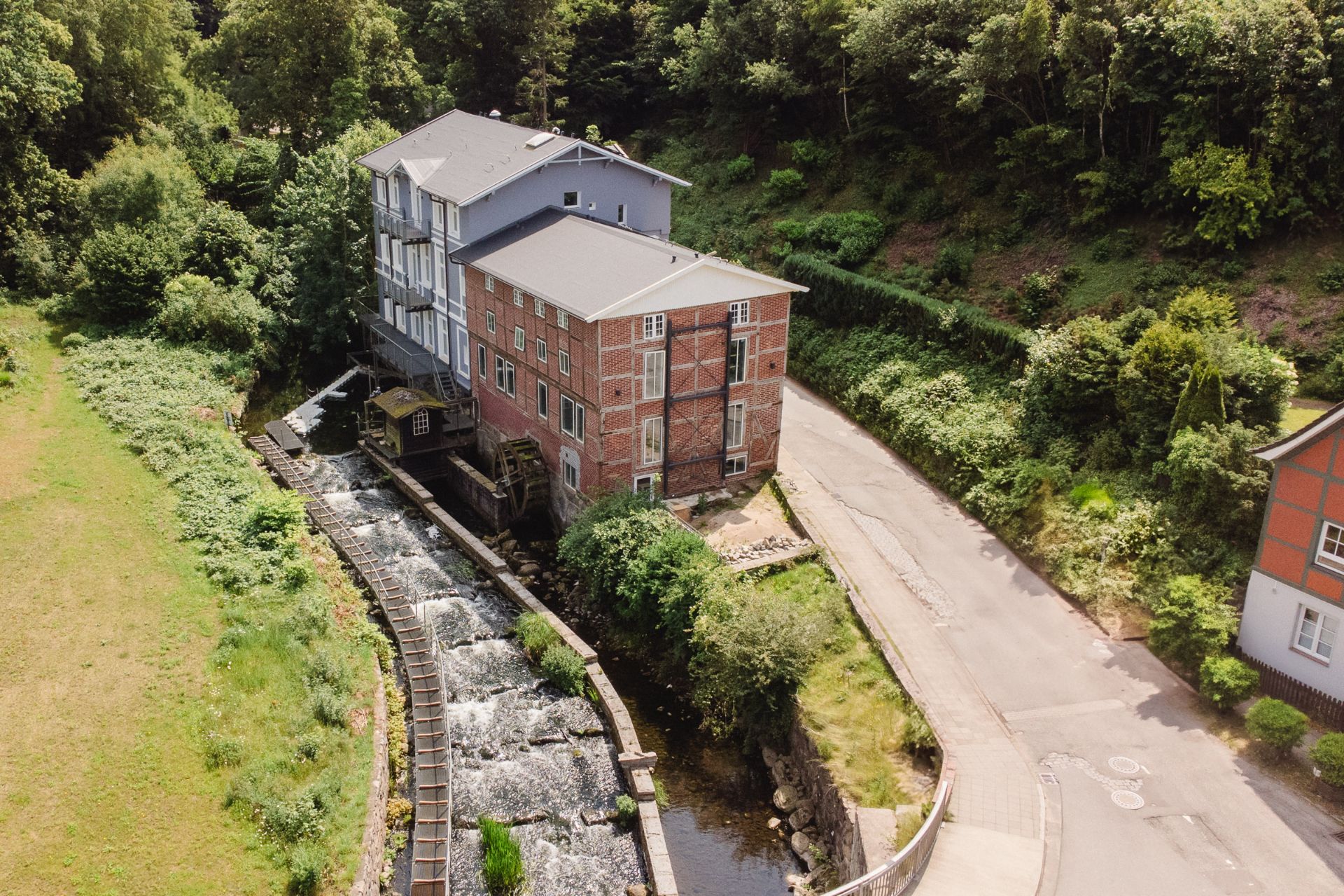 Großes Wohnhaus aus Backstein an einem Bach mit Wasserrad, umgeben von Bäumen und einer Landstraße.