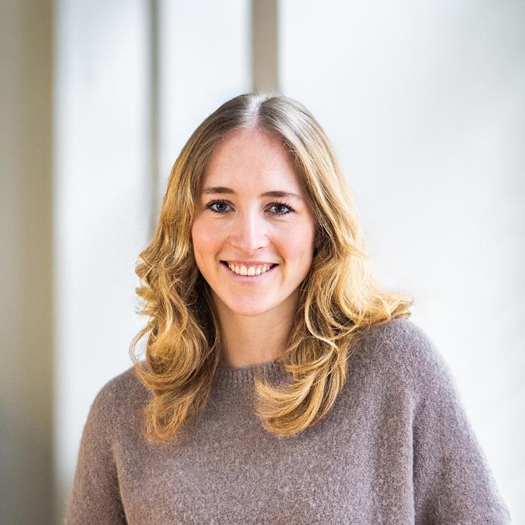 Junge Frau mit langen blonden Haaren steht in einem hellen Raum vor Fenstern.