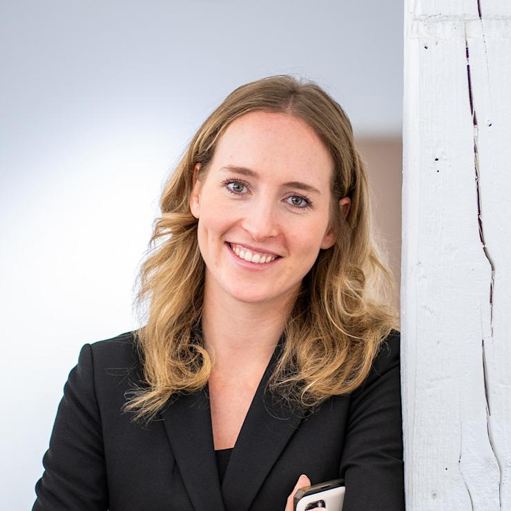 Junge Frau mit langen blonden Haaren steht in einem hellen Raum vor Fenstern.