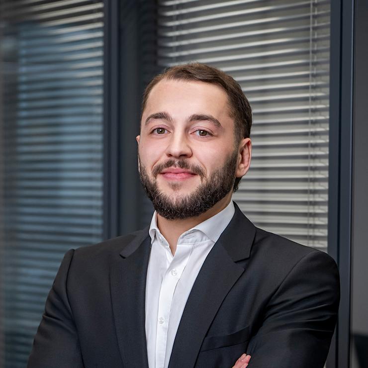 Mann im Anzug steht mit verschränkten Armen vor Fensterjalousien.