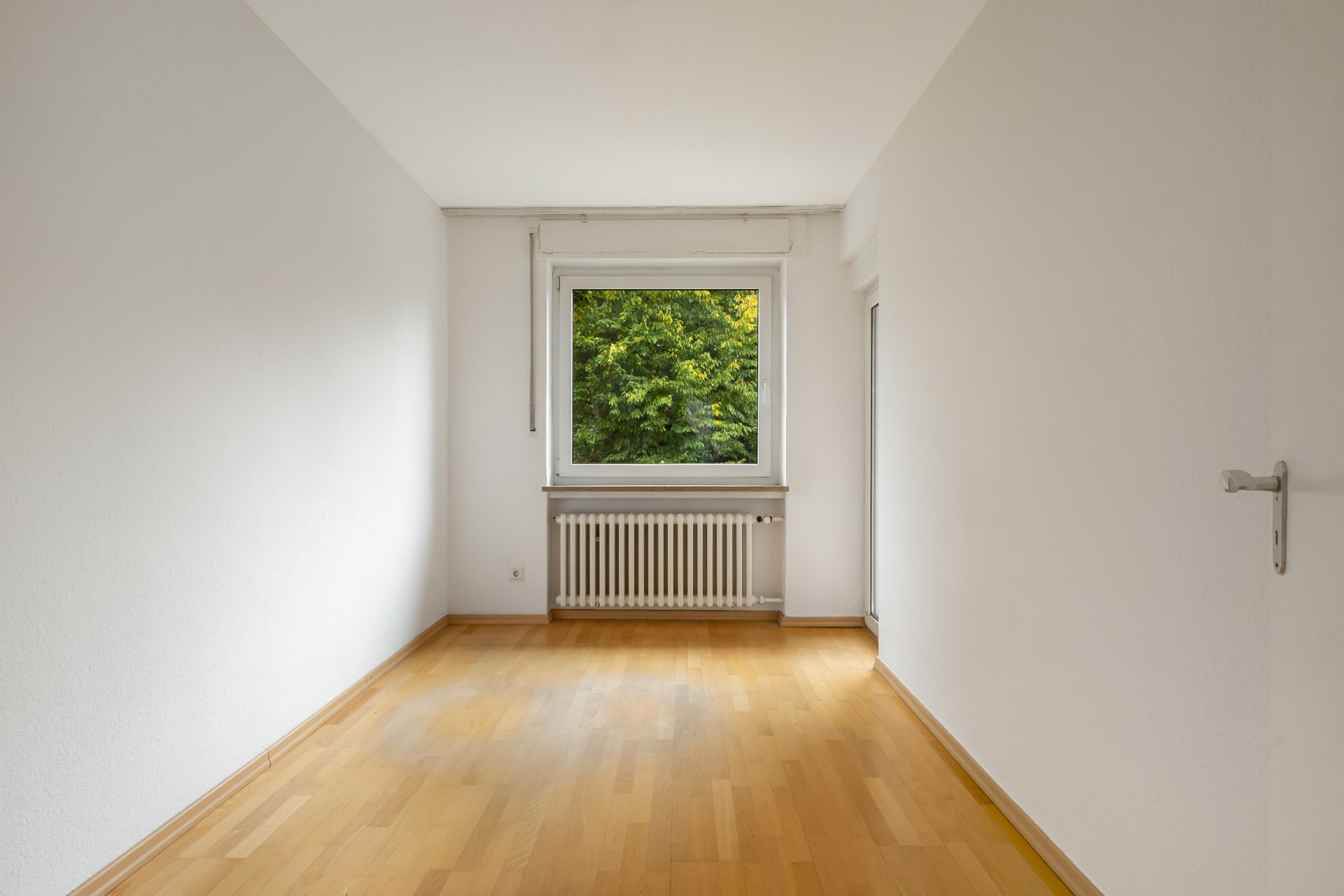 Leerer Raum mit Holzfußboden, weißer Wand, Fenster und Heizung darunter.