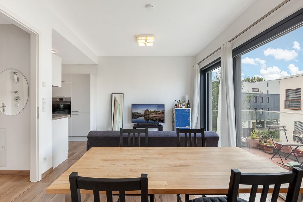 Helles Wohnzimmer mit Esstisch, Sofa, Fernseher und Zugang zu einem Balkon mit Pflanzen.